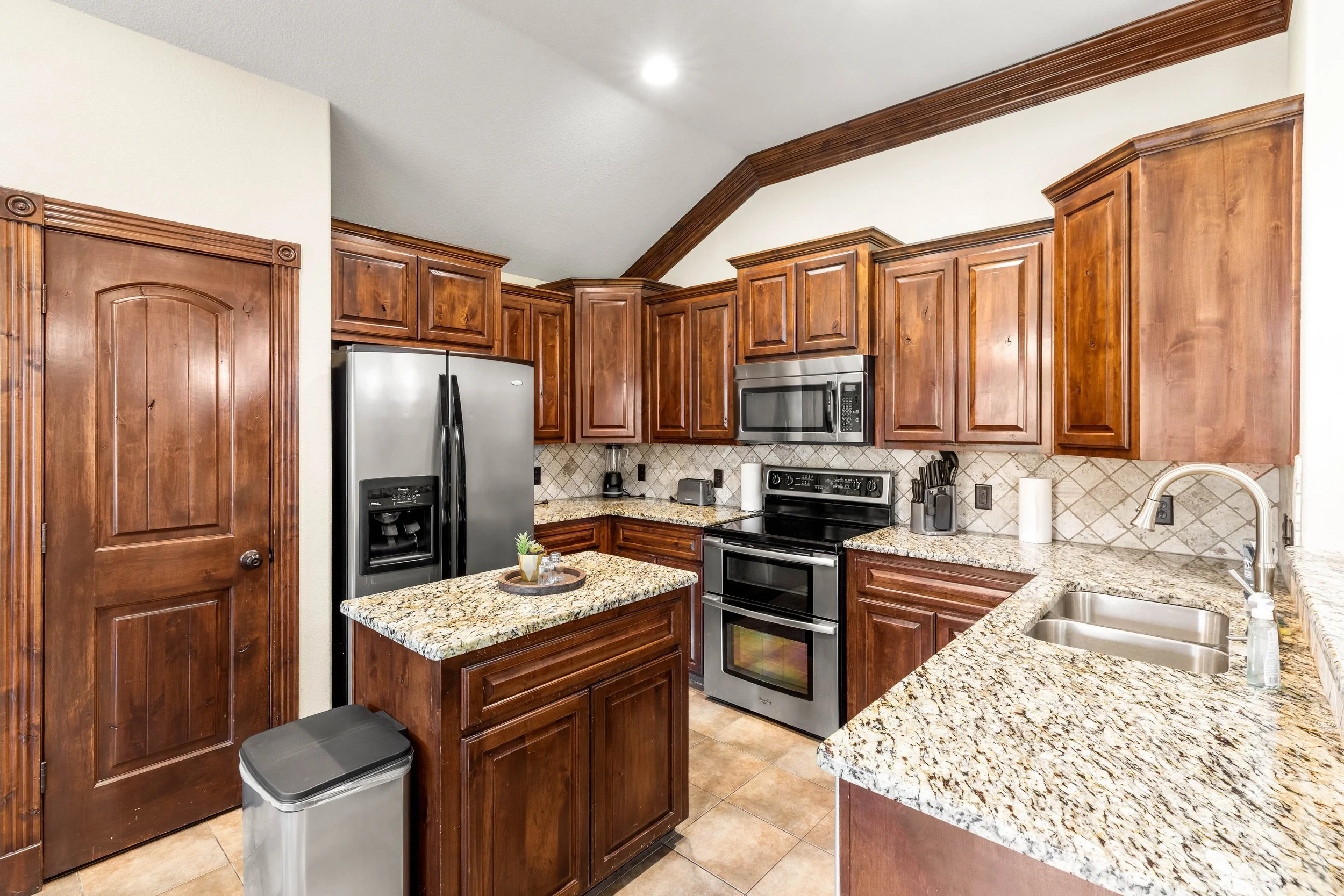Kitchen with cherry wood cabinetry, granite countertops, stainless steel appliances including a refrigerator and microwave, tiled backsplash, and stainless steel double sink.