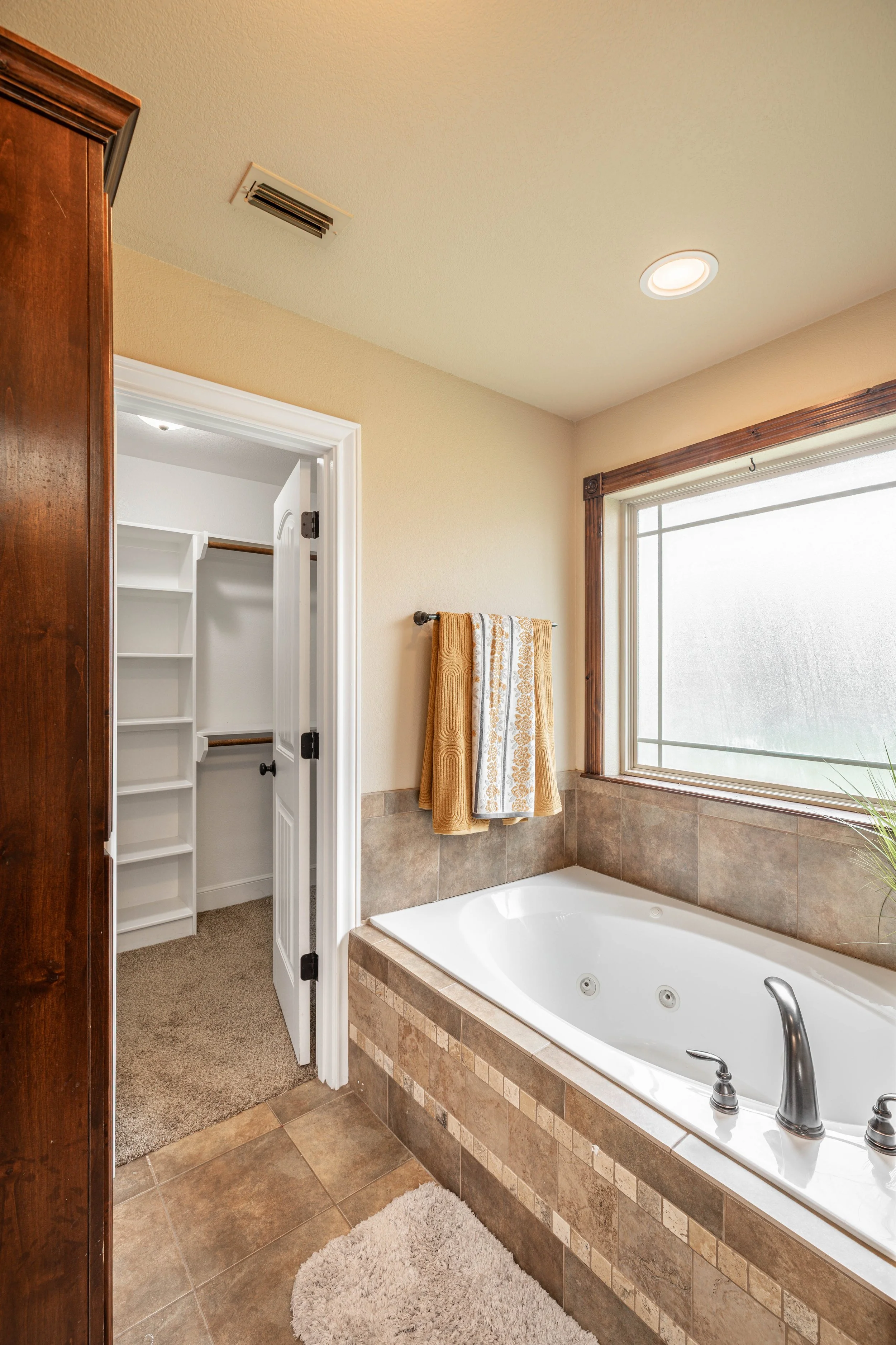 Bathroom with a built-in bathtub, a window with wooden trim, a towel rack with towels, and a closet with shelves visible through an open door. The room features beige walls, tiled surround for the tub, and a soft plush rug on a tiled floor.