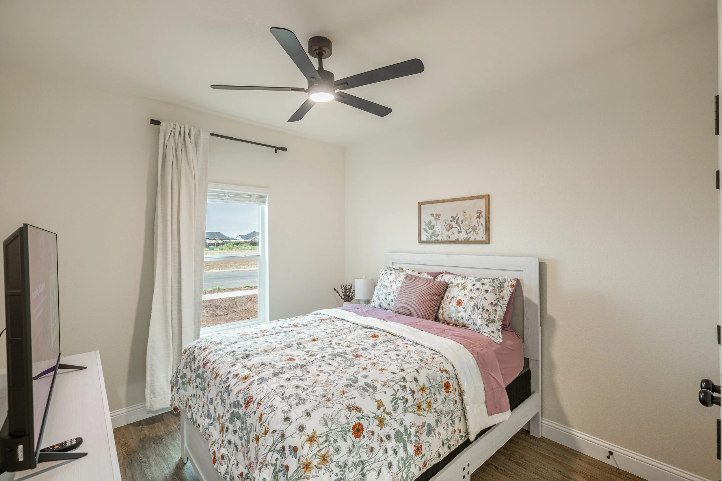 A bedroom with a white bed frame and floral bedding, a window with white curtains, a framed botanical print on the wall, a black ceiling fan with a light, a TV on a white stand, and hardwood floors.