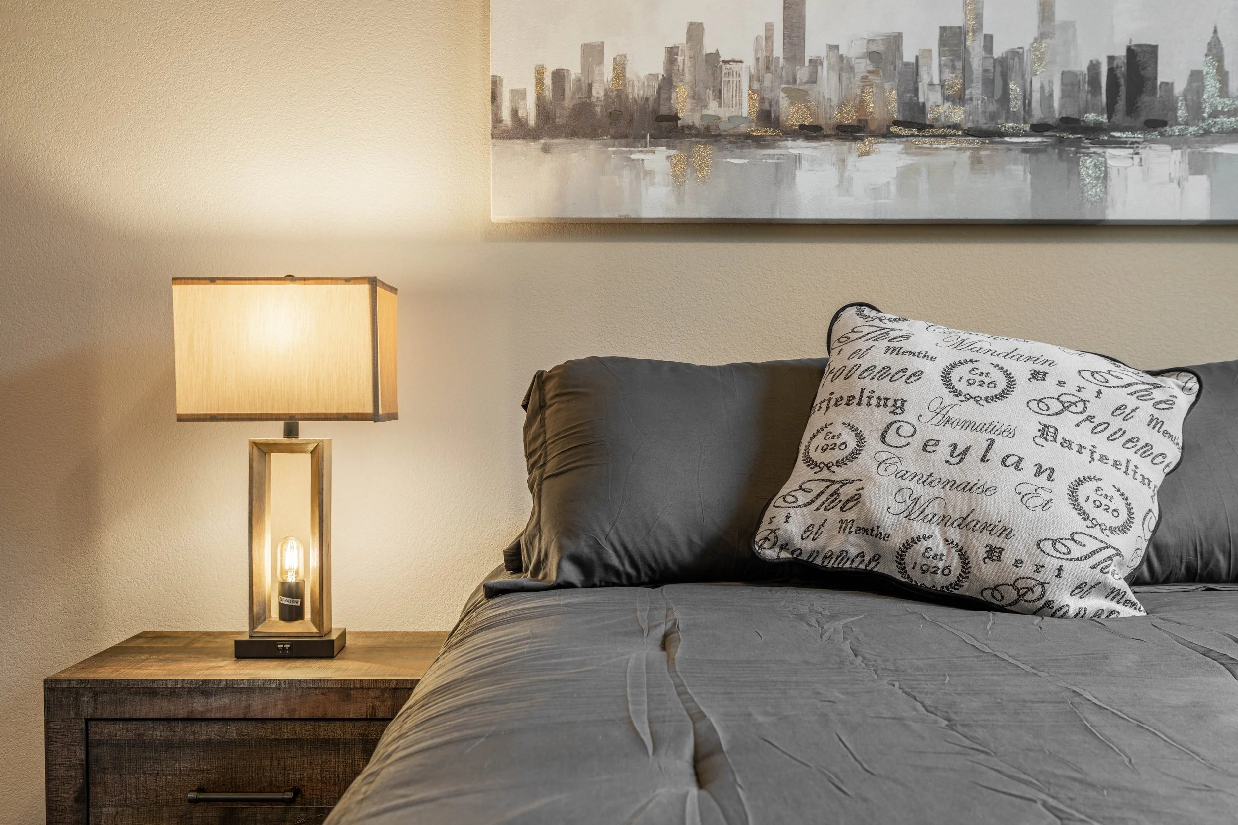 A bedside lamp on a wooden nightstand next to a bed with a decorative pillow and gray bedding, with a cityscape painting hanging on the wall above the bed.