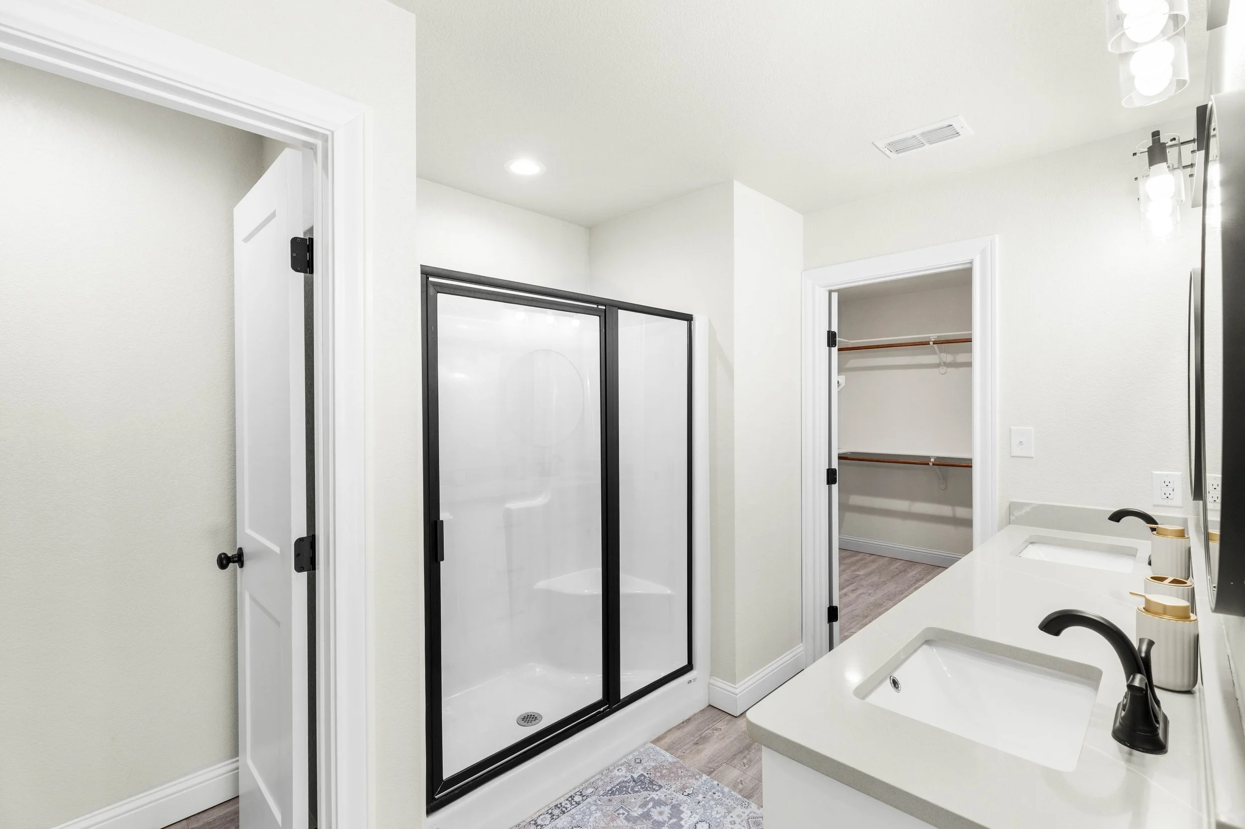 Bathroom with white walls, a white sink with a black faucet, a small mirror, a black framed glass shower enclosure, an open closet with a wooden rod, and ceiling lights.