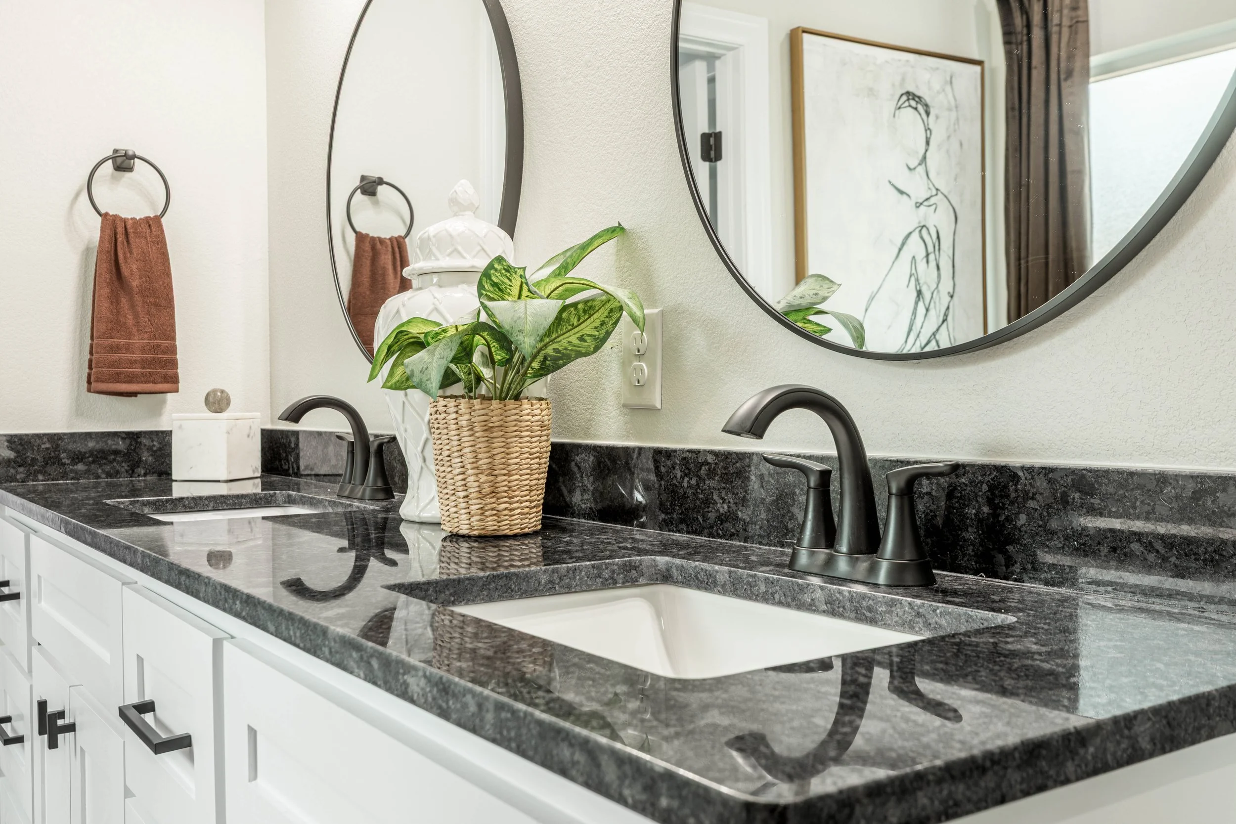 A bathroom bathroom countertop with a black granite surface, a white cabinet with black handles below, a bronze faucet, a green potted plant in a woven basket, a white decorative jar, a brown hand towel hanging on a black towel ring, and a mirror ref