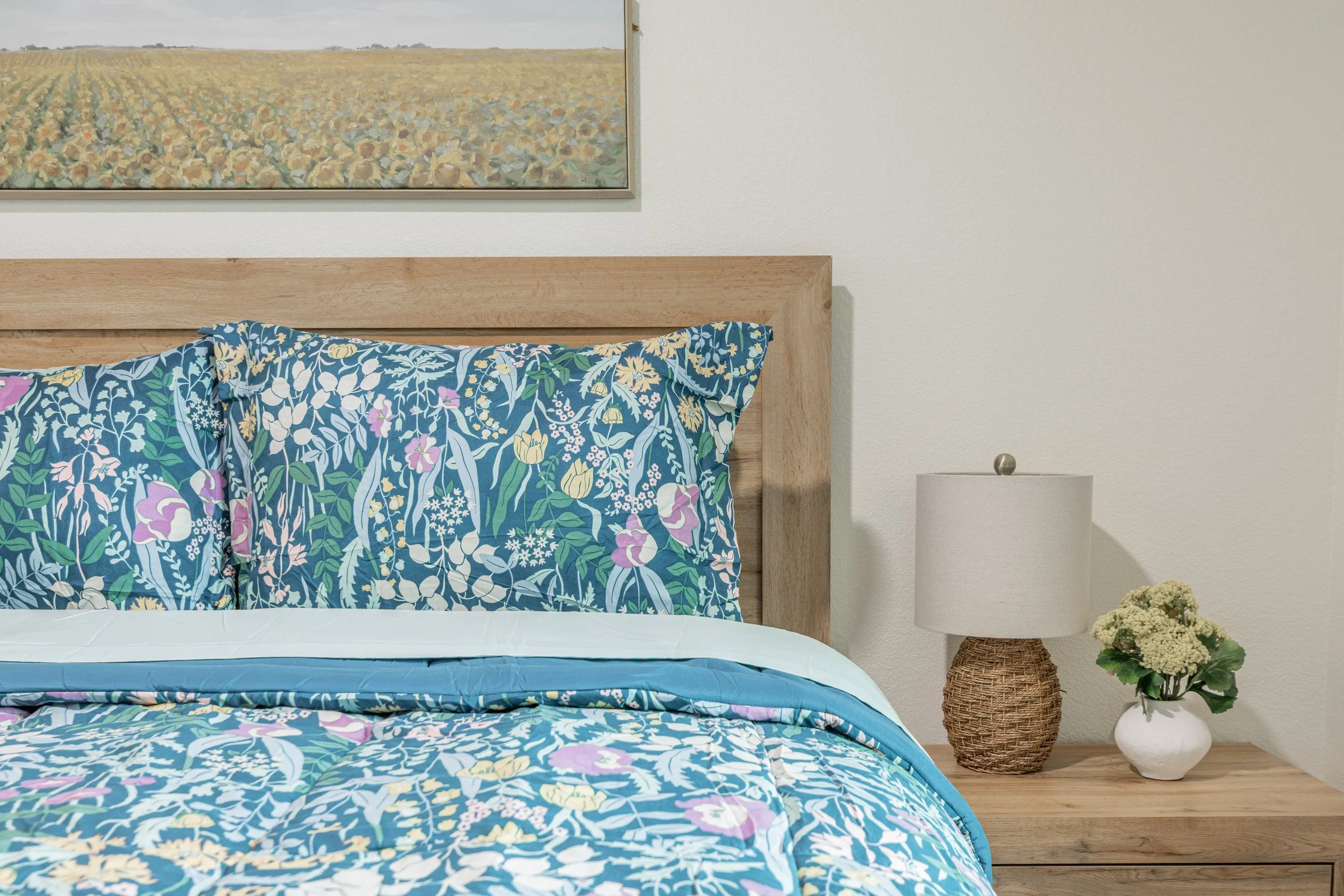 A bedroom with a wooden bed frame, blue floral bedspread, and matching pillowcases, a white wall, a framed landscape painting of a sunflower field, a white nightstand with a lamp, and a white vase with flowers.