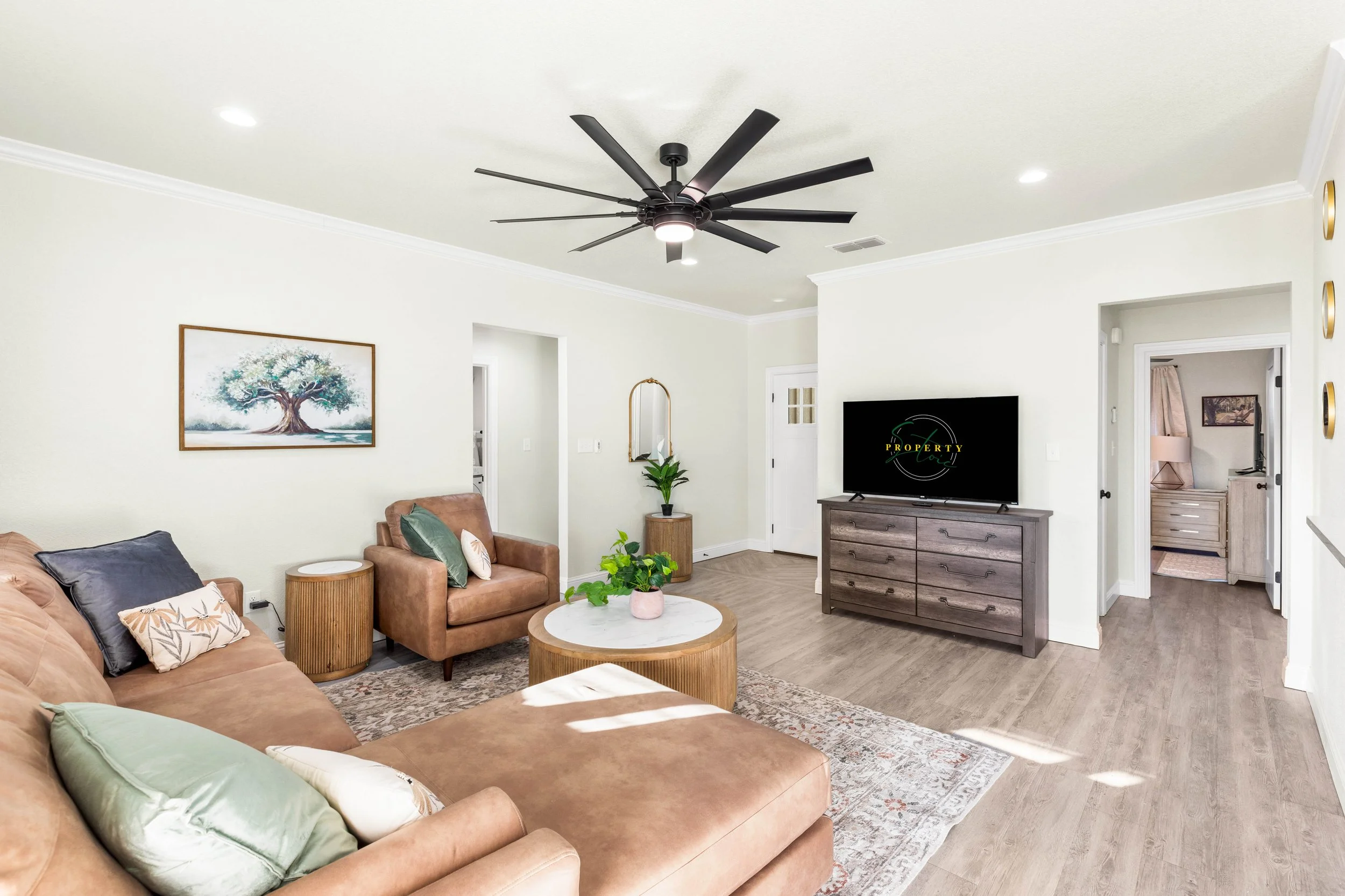 Living room with beige couch, green and patterned pillows, coffee tables with a pink plant, wall art, a TV on a wooden stand, a ceiling fan, and a doorway leading to another room.