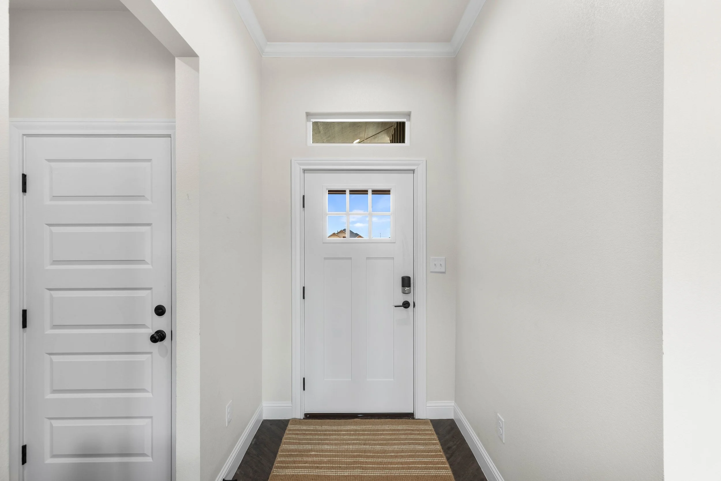 Entryway with white walls, a white front door with a window, a narrow high window above it, and a small window to the left. There is a striped rug on dark wood flooring.