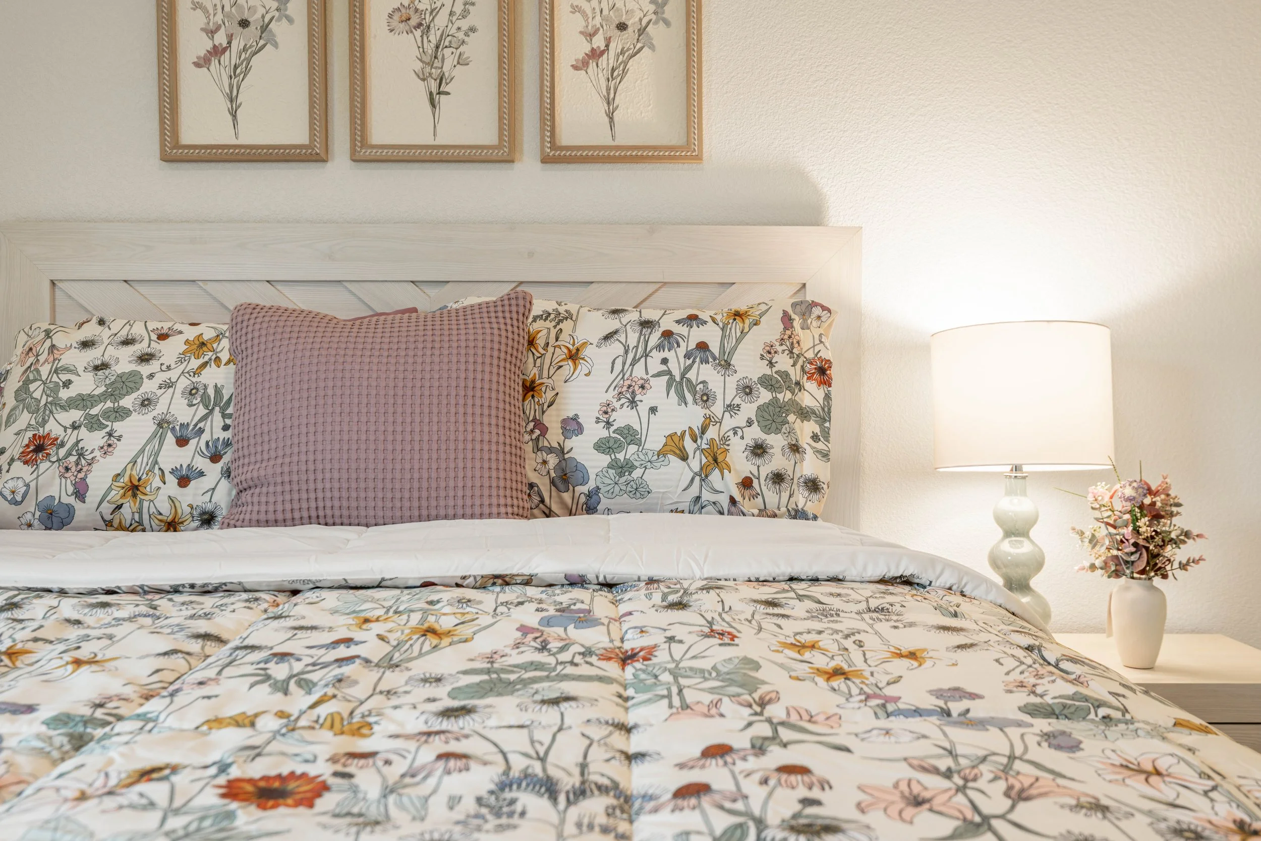 Close-up of a neatly made bed with floral-patterned bedding, a pink textured pillow, a white bedside table with a lamp, and framed botanical prints hanging on the beige wall behind.