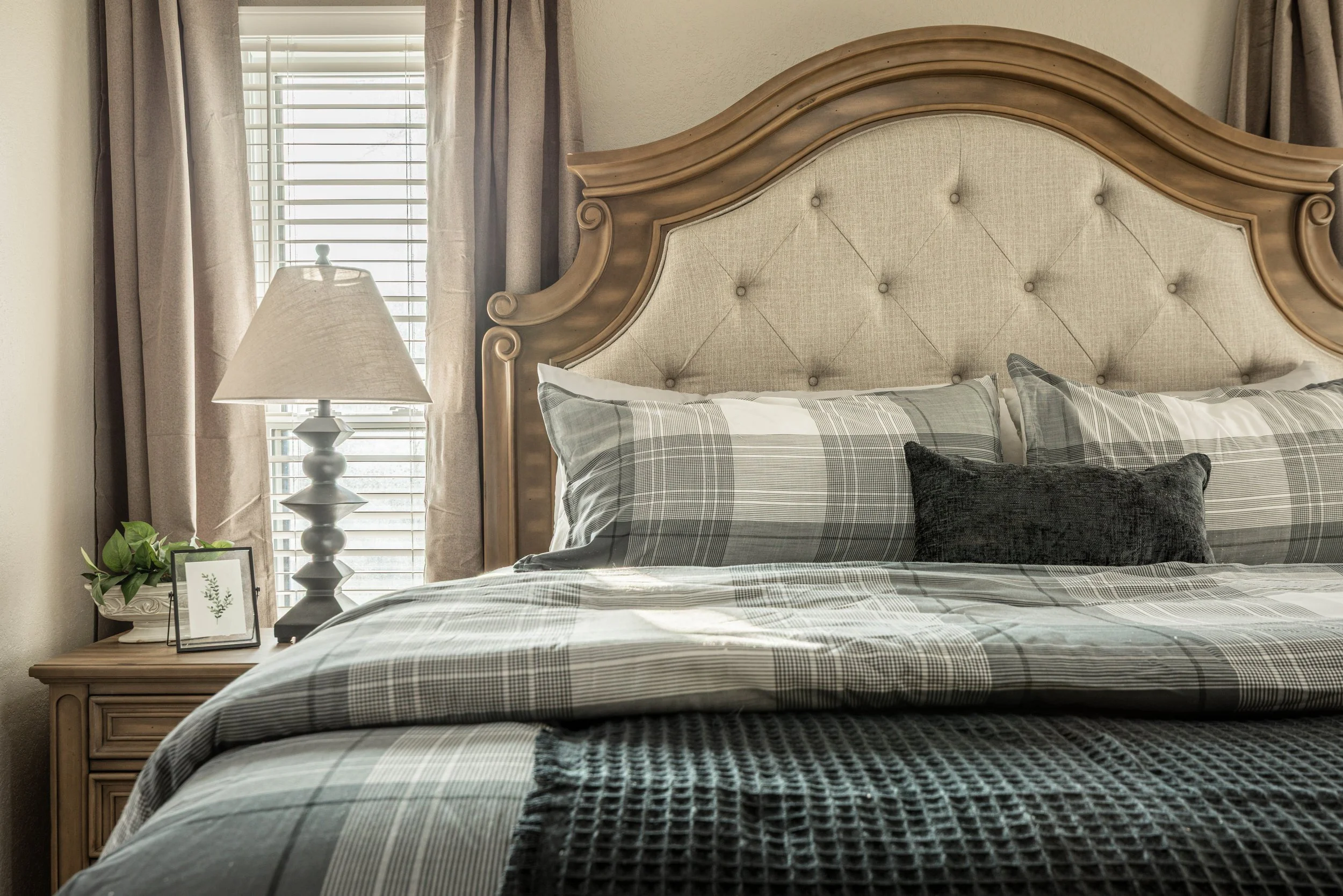 A bedroom with a large bed featuring a tufted beige headboard with wooden accents, plaid bedding, and black throw pillows. Beside the bed is a wooden nightstand with a white table lamp, a small framed picture of greenery, and a plant. The room has be