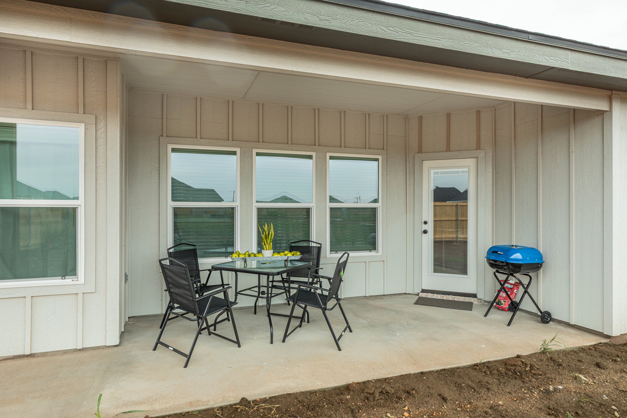 Back porch area of a house with patio furniture, a blue barbecue grill, and large windows. There are some green apples and plants on the table.