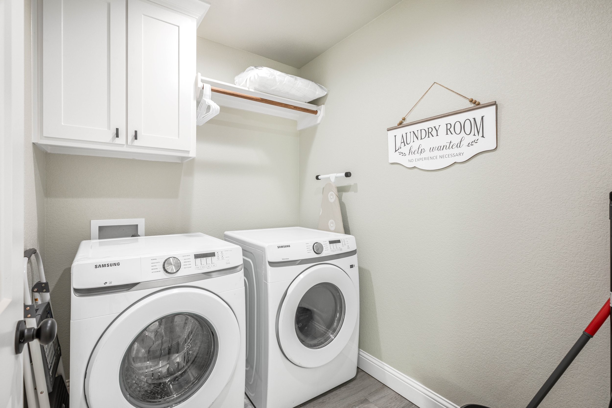 A laundry room with a white cabinet, a shelf with a pillow, a sign that says "Laundry Room help wanted, no experience necessary," and two white Samsung washing machines.