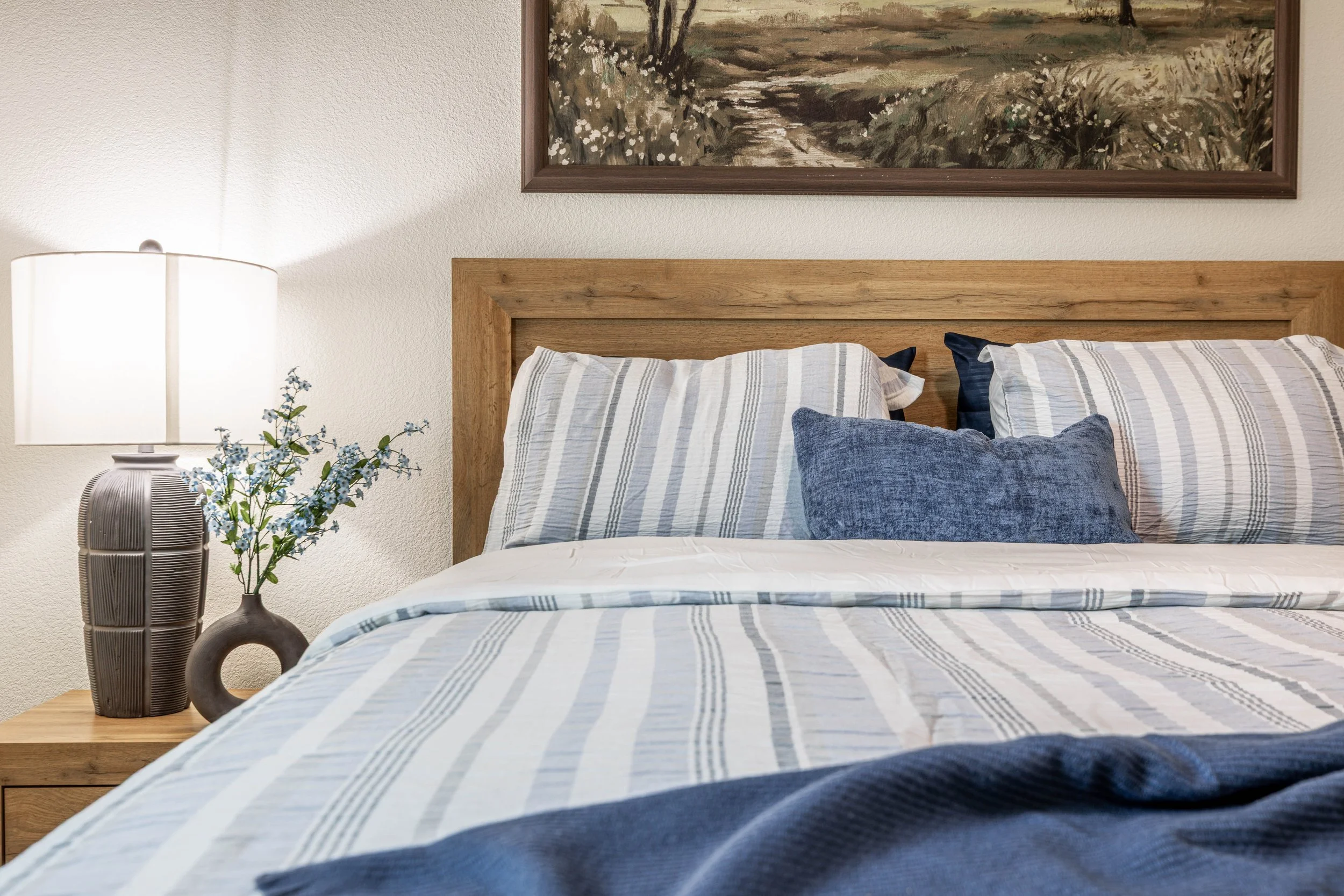 A neatly made bed with a wooden headboard, striped bed linens, and multiple pillows in shades of blue and white. To the left, a bedside table holds a decorative lamp and a small vase with flowers. Above the bed, a landscape painting hangs on the wall