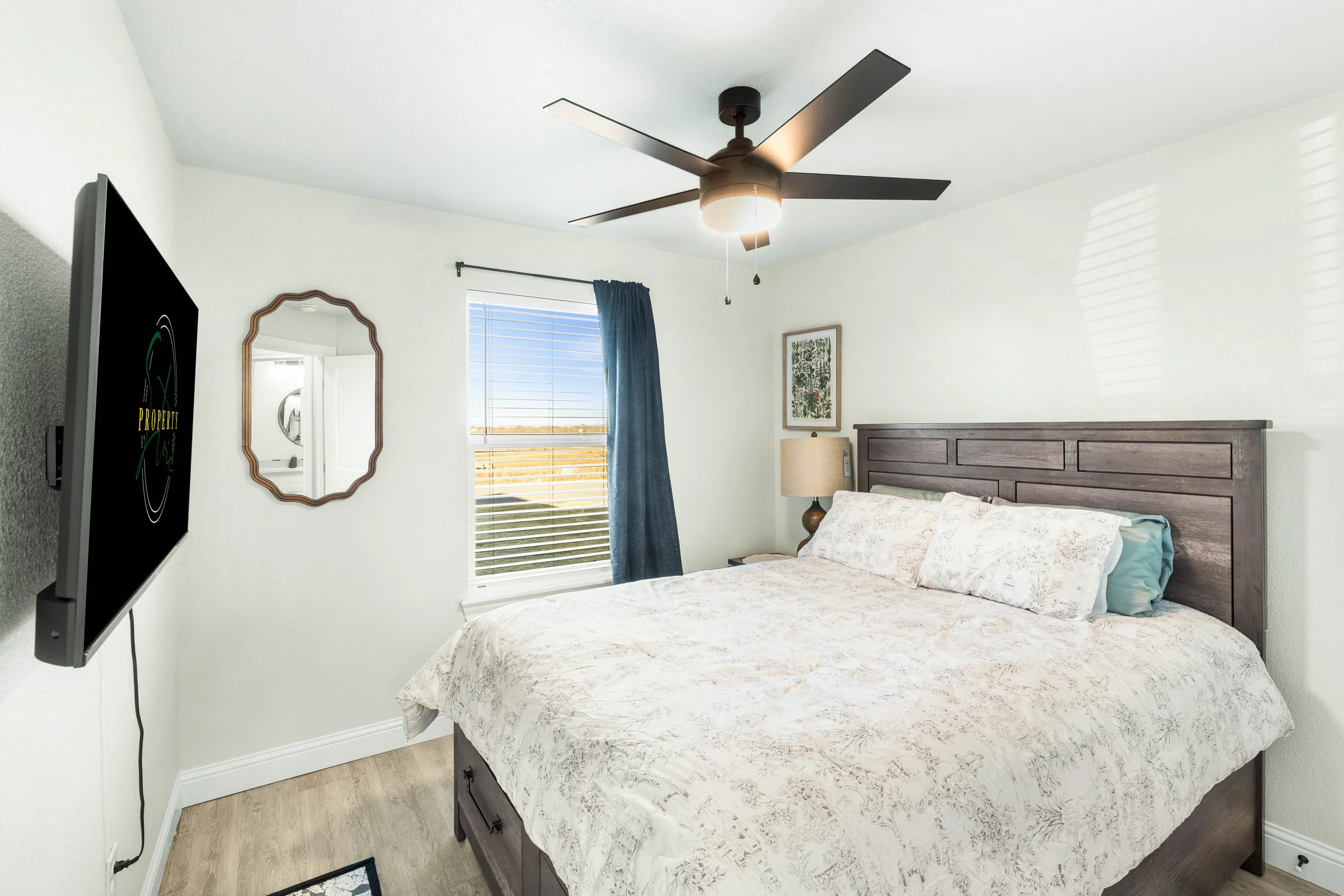 A bedroom with a bed, wooden headboard, nightstand, lamp, wall art, window with blue curtains, a ceiling fan, and a wall-mounted TV.
