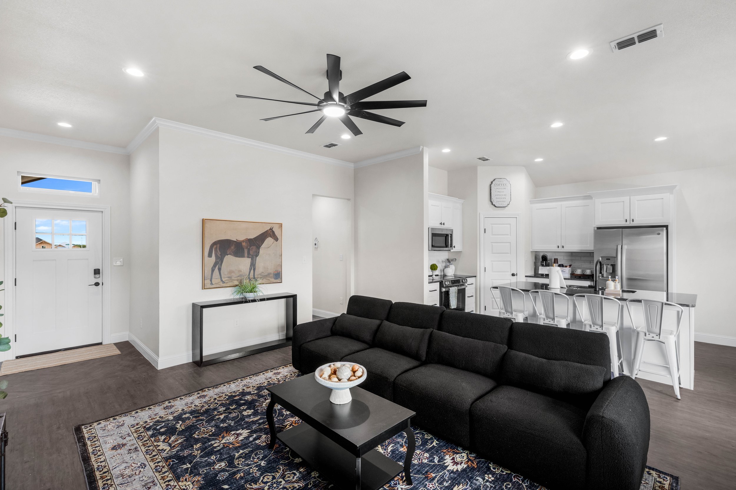 Living room with white walls, black sofa, colorful area rug, black coffee table with decorative bowl, white kitchen with stainless steel appliances, white dining table with chairs, recessed lighting, ceiling fan, artwork of a horse, front door with w