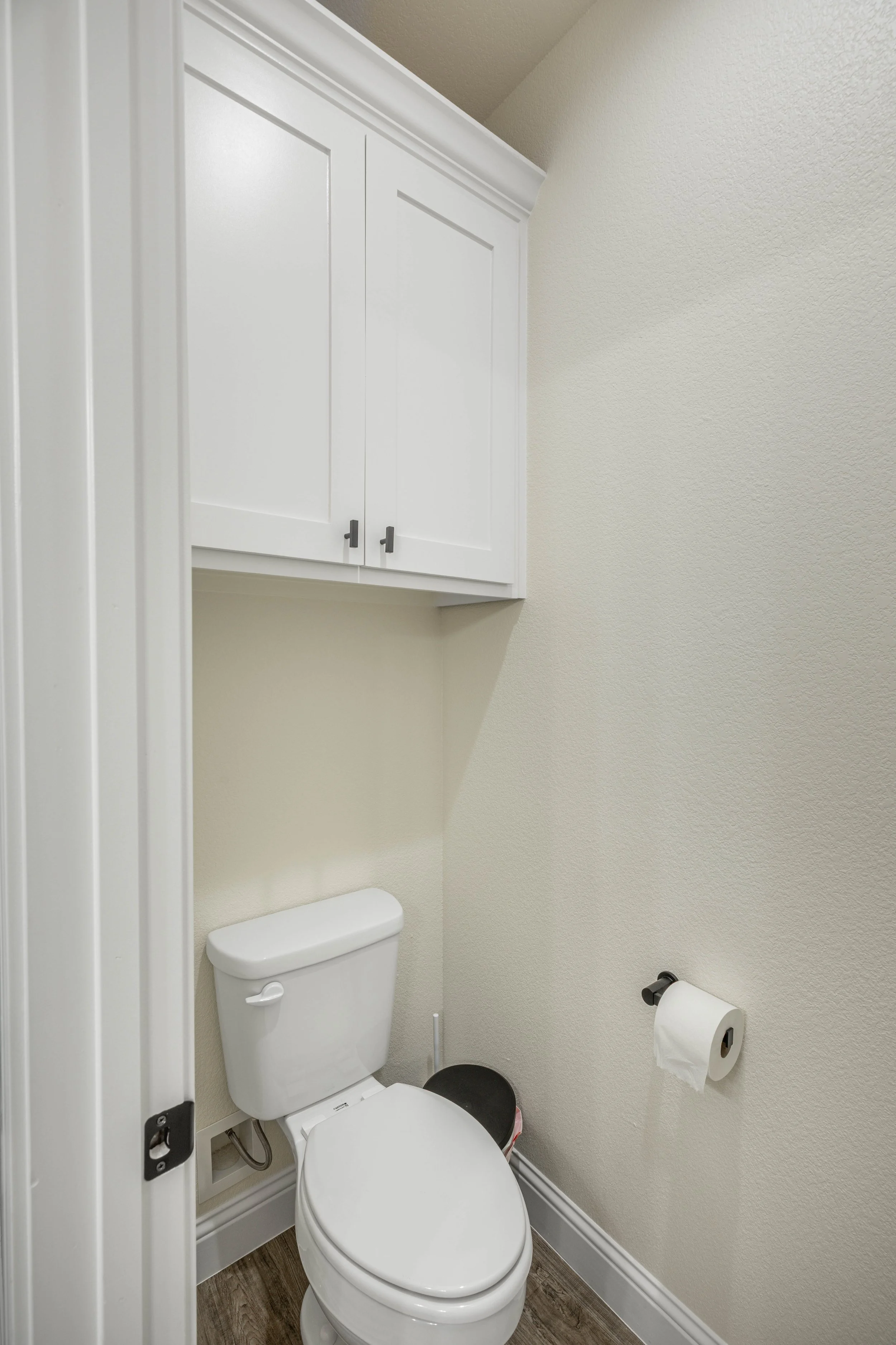 A small bathroom with a white toilet, a wall-mounted cabinet above, a toilet paper roll on the wall, and a dark brown wood floor.
