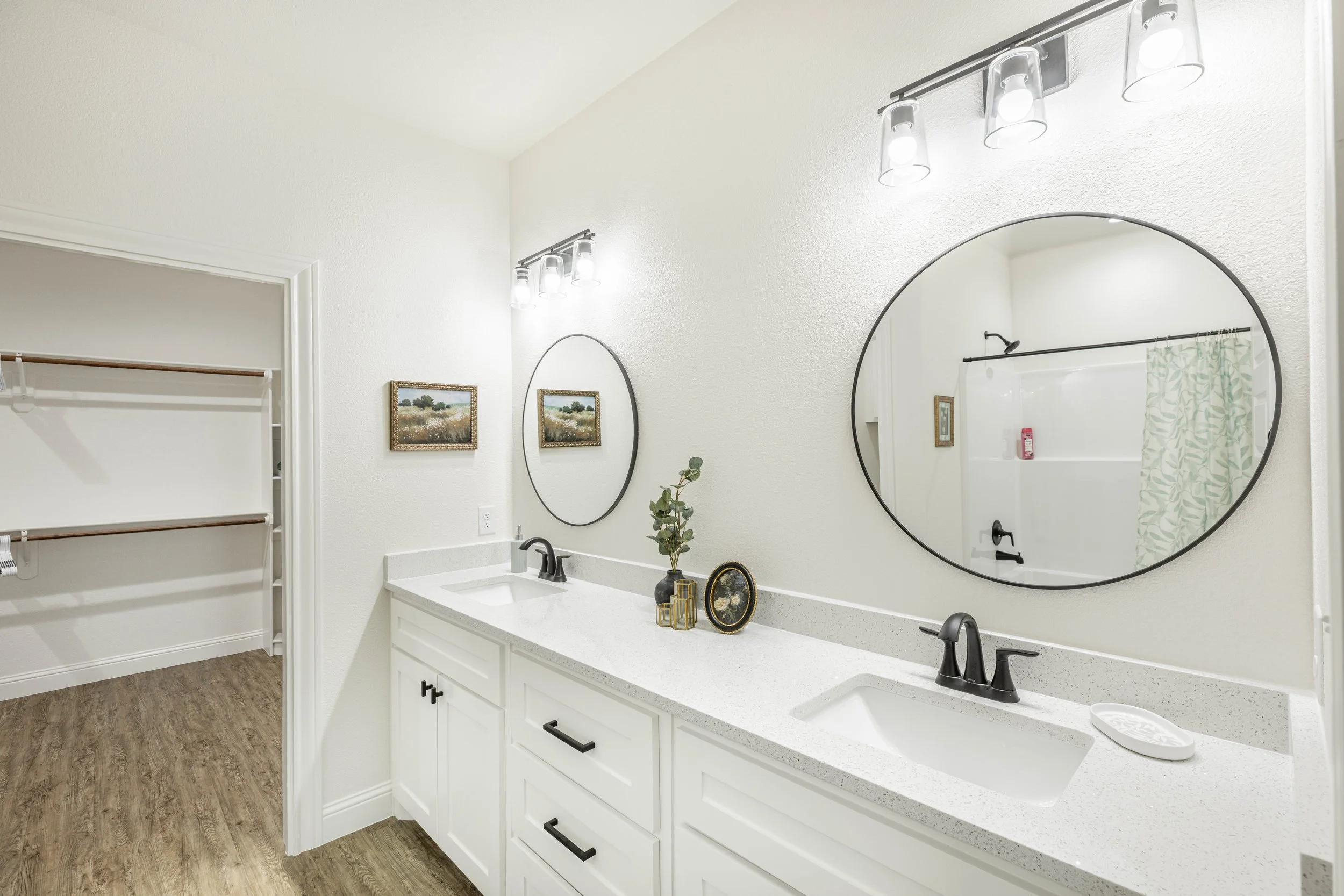 Bathroom with white vanity, round mirrors, black fixtures, decorative items, and framed landscape artwork. Part of a shower with green leaf patterned curtain is visible.