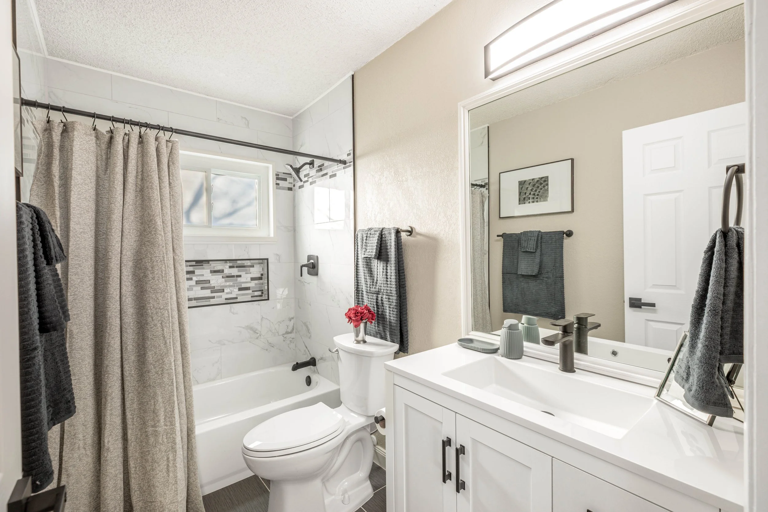 A modern bathroom with a shower area separated by a beige curtain, a toilet, a white vanity with a mirror, gray towels, a small flower pot, and a window with daylight.