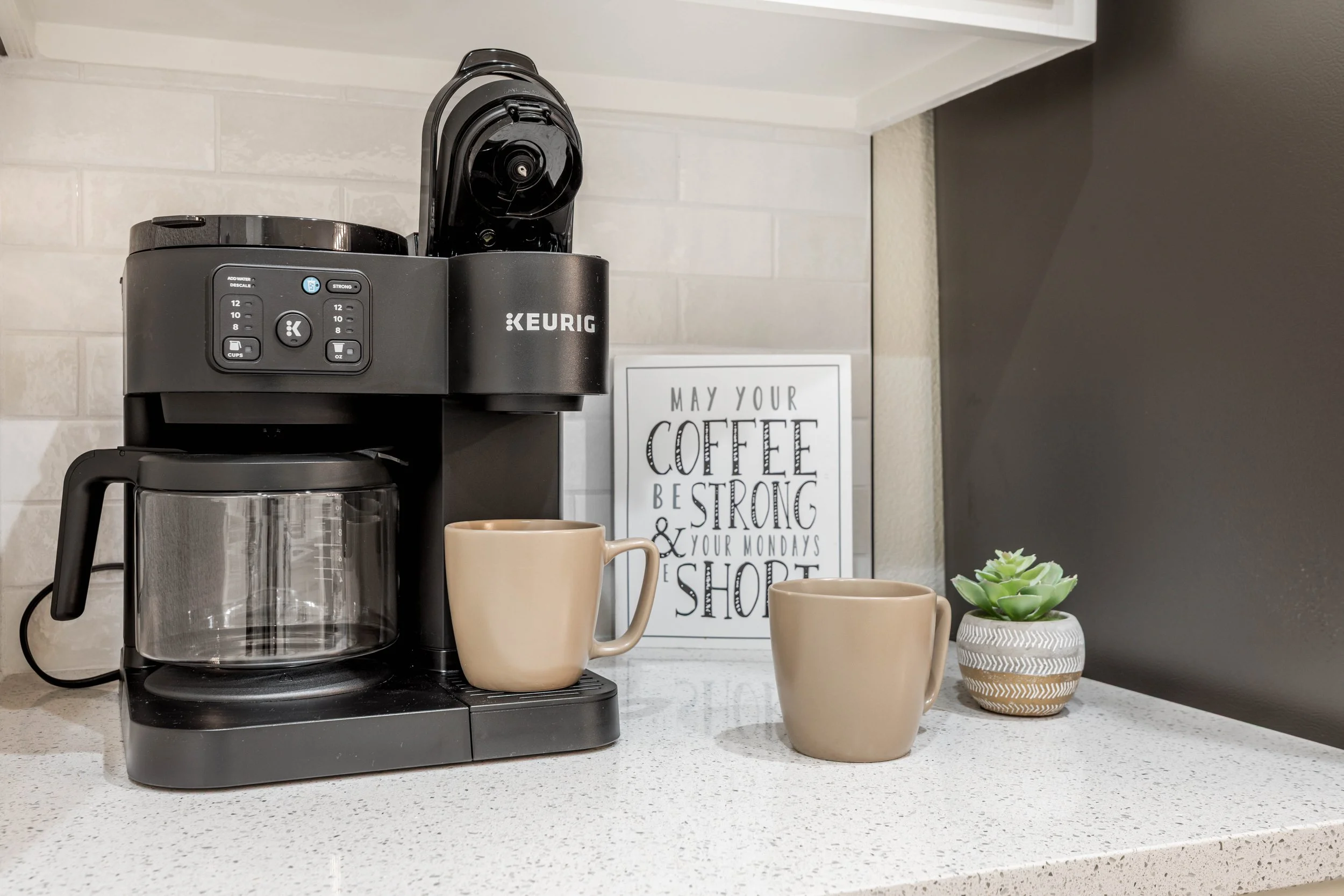 Black coffee maker on a kitchen countertop with two beige mugs, a small potted succulent, and a framed sign reading 'May your coffee be strong and your Monday be short.'