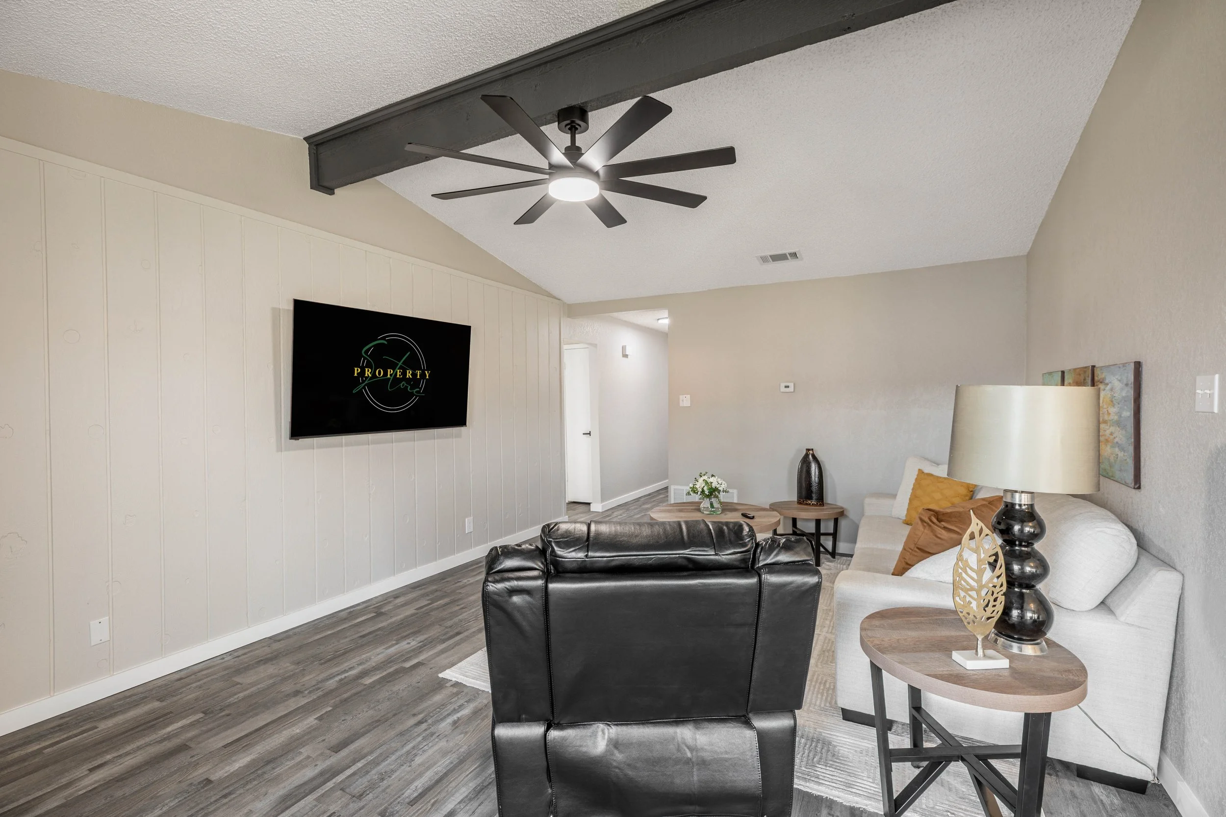 Living room with a ceiling fan, wall-mounted TV, beige walls, gray flooring, cream sofa with mustard and beige pillows, side table with decorative items, and a small table with a floral arrangement.