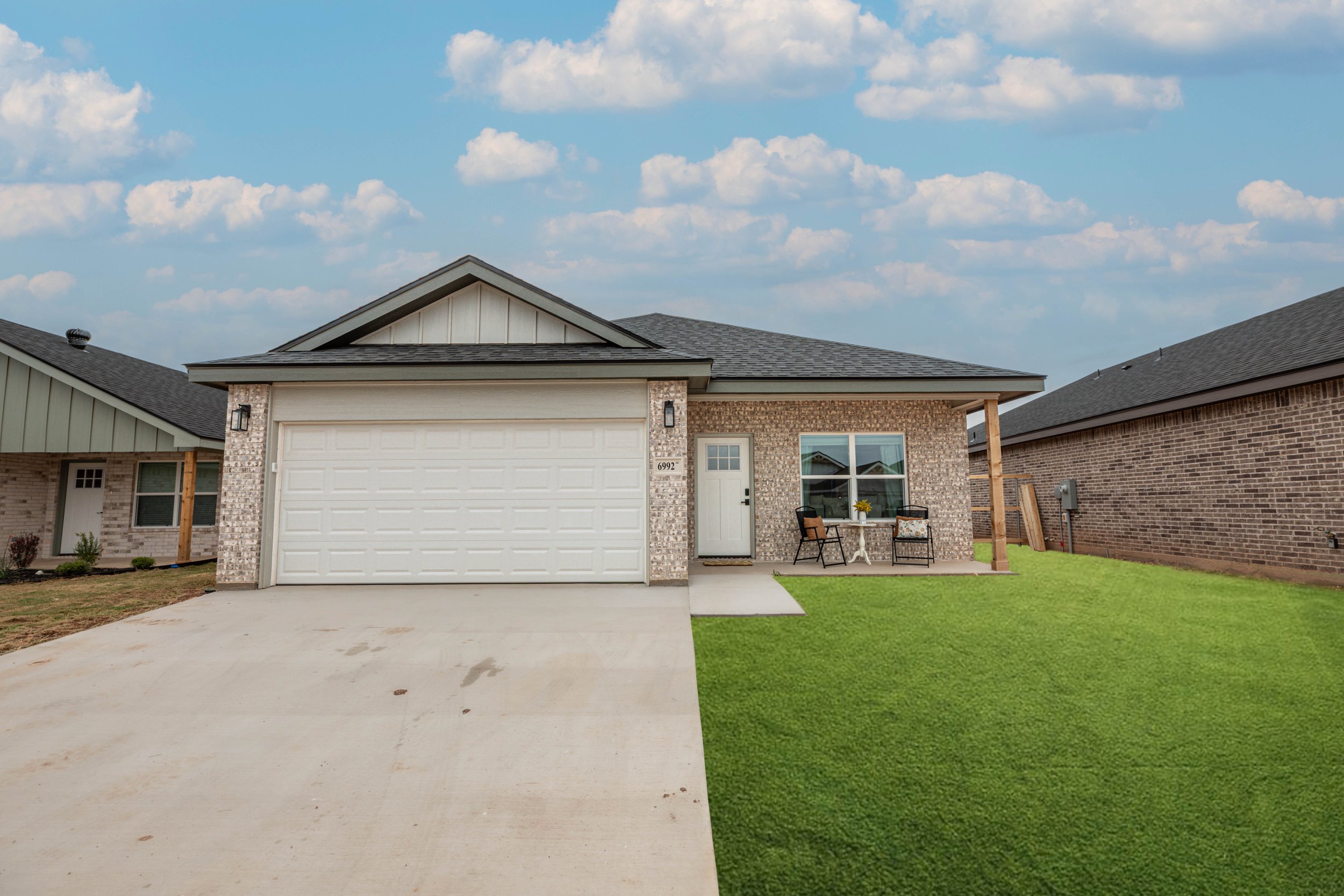 A newly built brick house with a white garage door, front porch with two chairs and a small round table, green lawn, neighboring houses on either side, and a partly cloudy sky overhead.