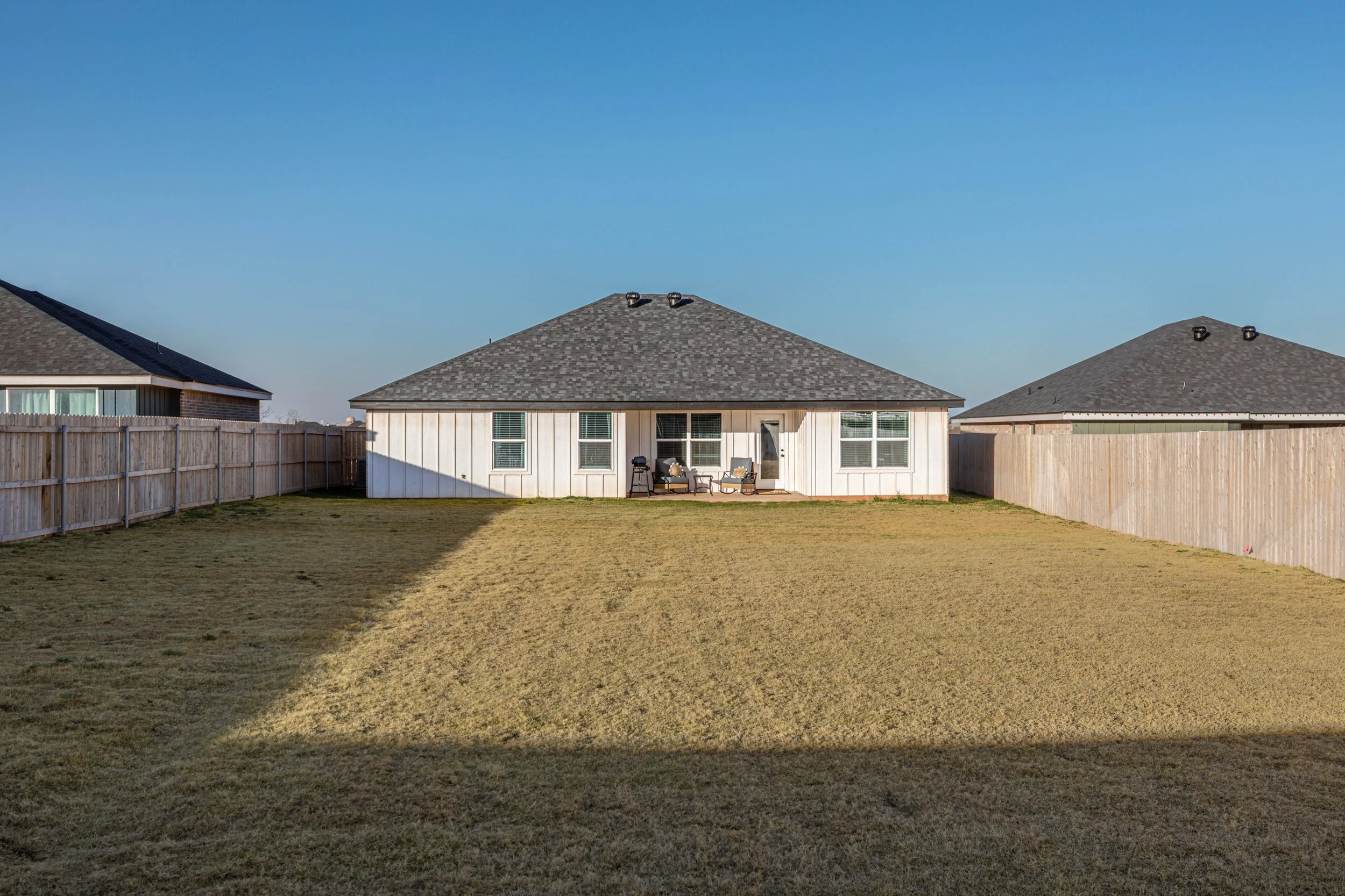 Backyard with a grassy lawn, wooden privacy fence, and a single-story house with a gray shingle roof, patio furniture, and a barbecue grill.