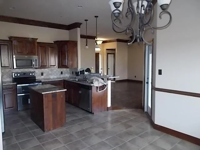 Empty kitchen with dark wooden cabinets, stainless steel appliances, and a tiled floor, with an open living area and a chandelier overhead.