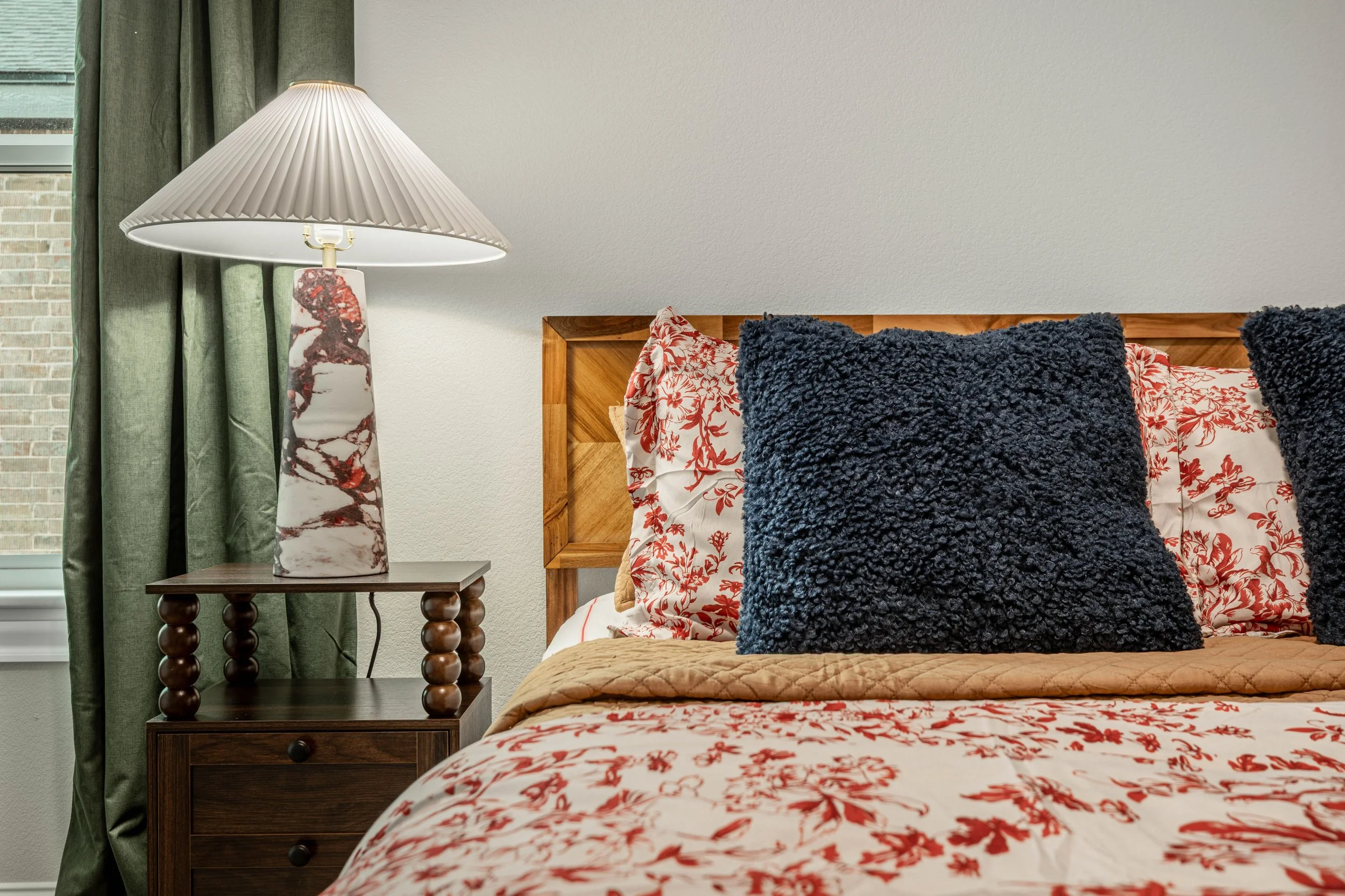 A bedroom with a wooden bed frame, floral bedspread, and decorative pillows including dark blue textured pillows. A side table with a marble-patterned lamp and green curtains near a window are also visible.