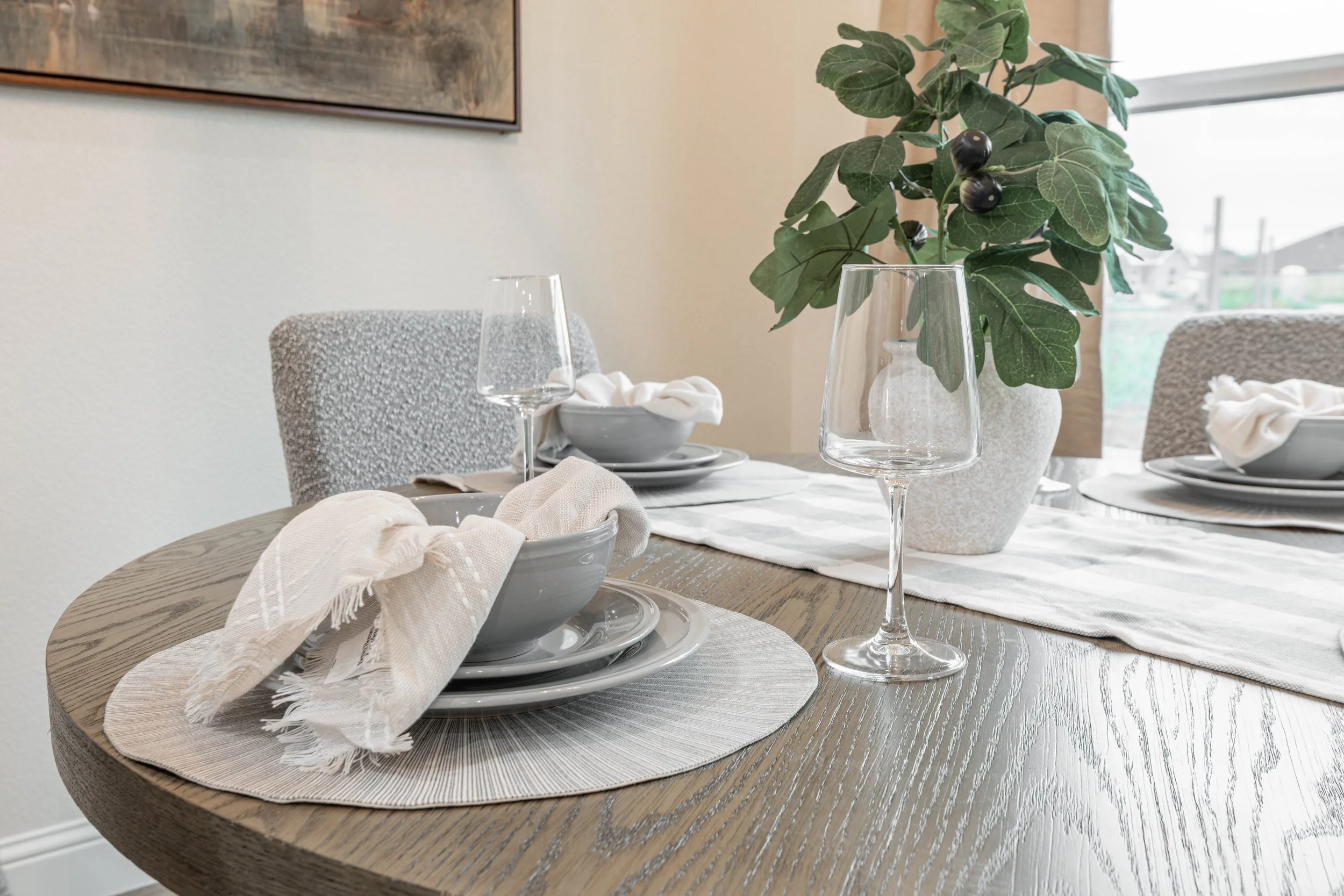 A dining table set with white plates, bowls, beige napkins, wine glasses, and a large potted green plant with dark berries as a centerpiece. There are upholstered chairs and a window in the background.