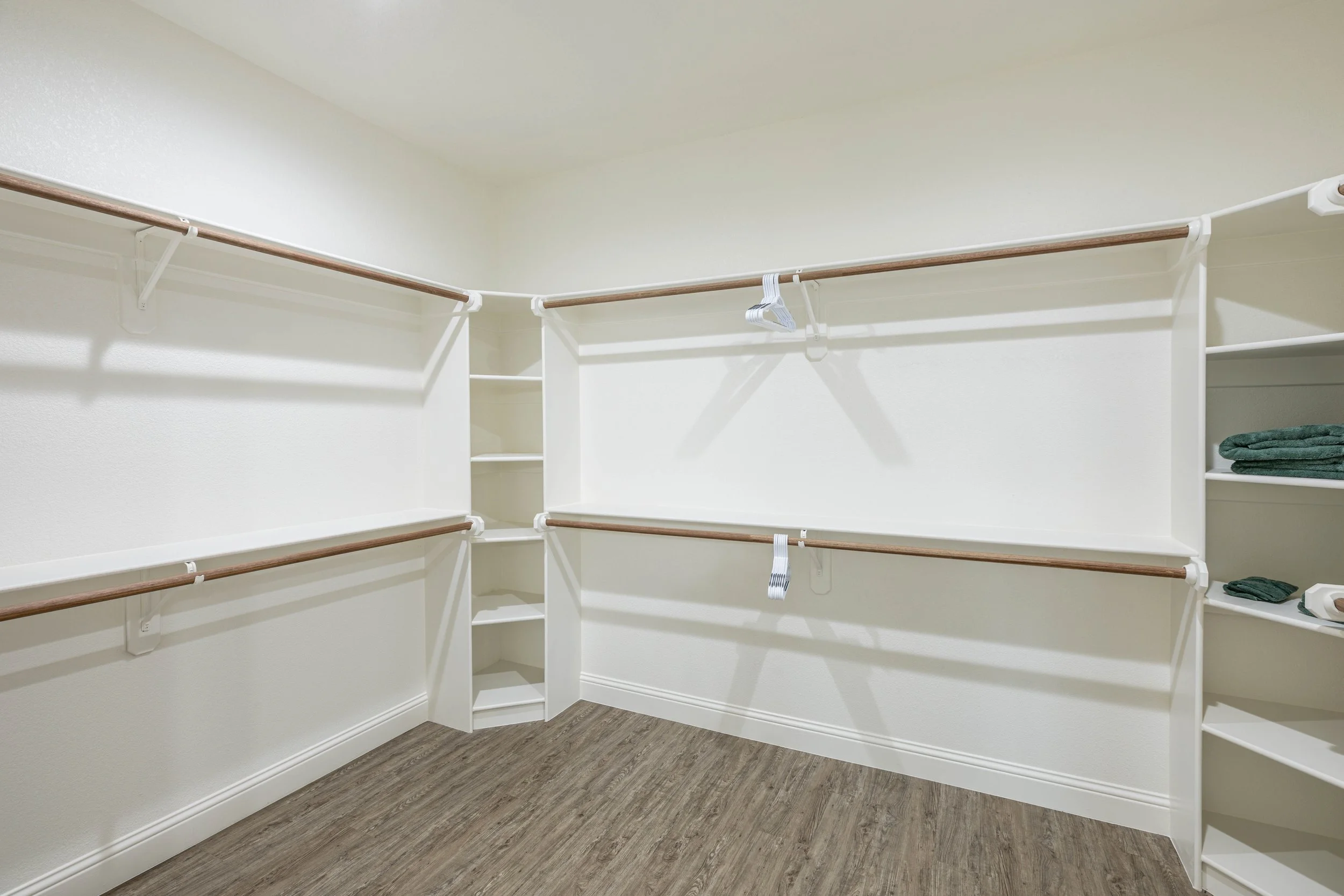 Empty walk-in closet with white shelving, wood hanging rods, and folded towels, with wood flooring.