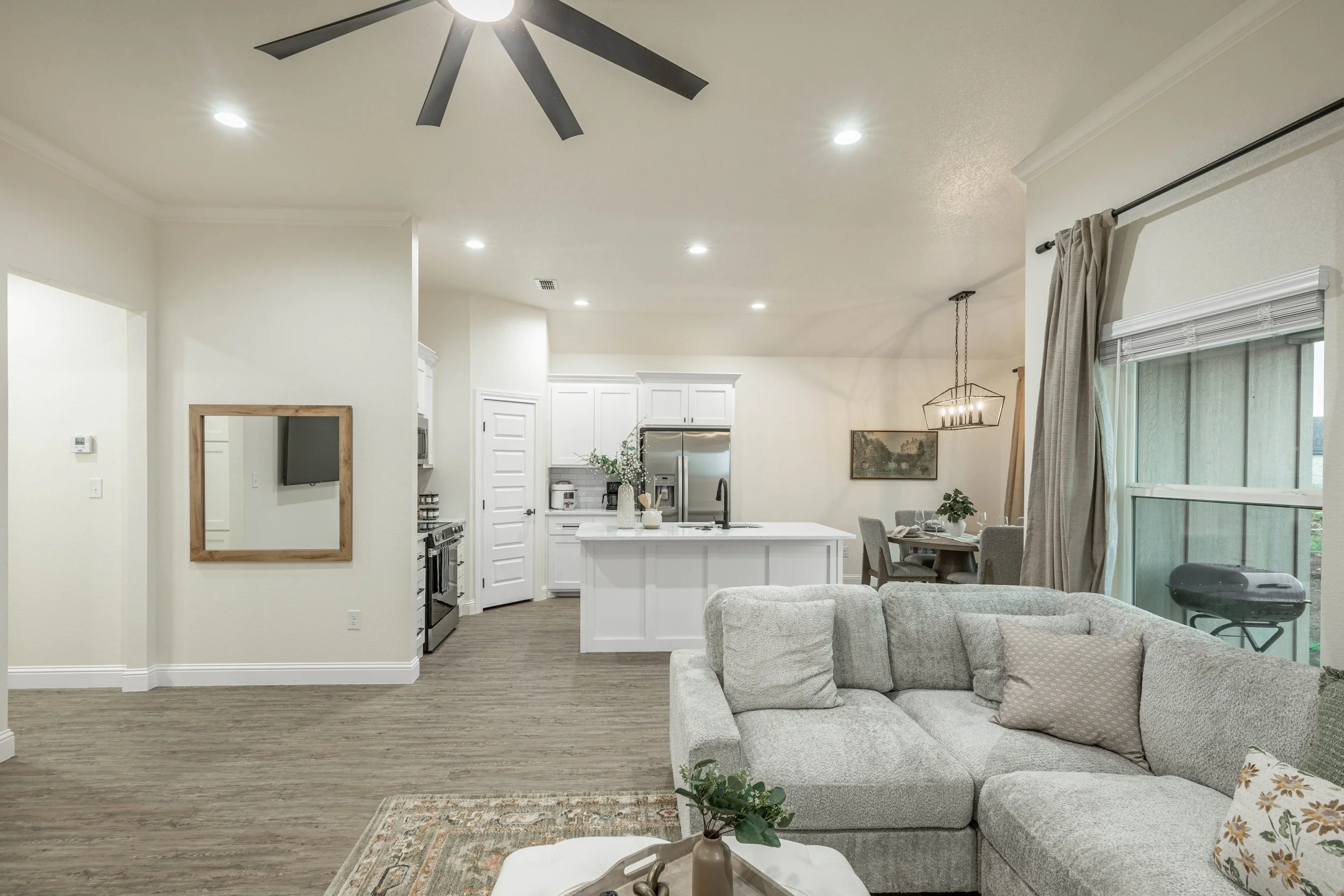 Open-concept living room and kitchen with white cabinets, stainless steel refrigerator, dining table with chairs, sofa, and large window with curtains.