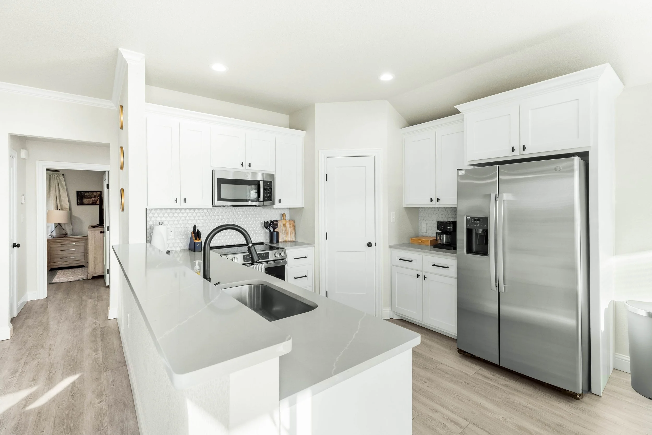 Modern white kitchen with stainless steel appliances, white cabinets, a large island, and light wood flooring.