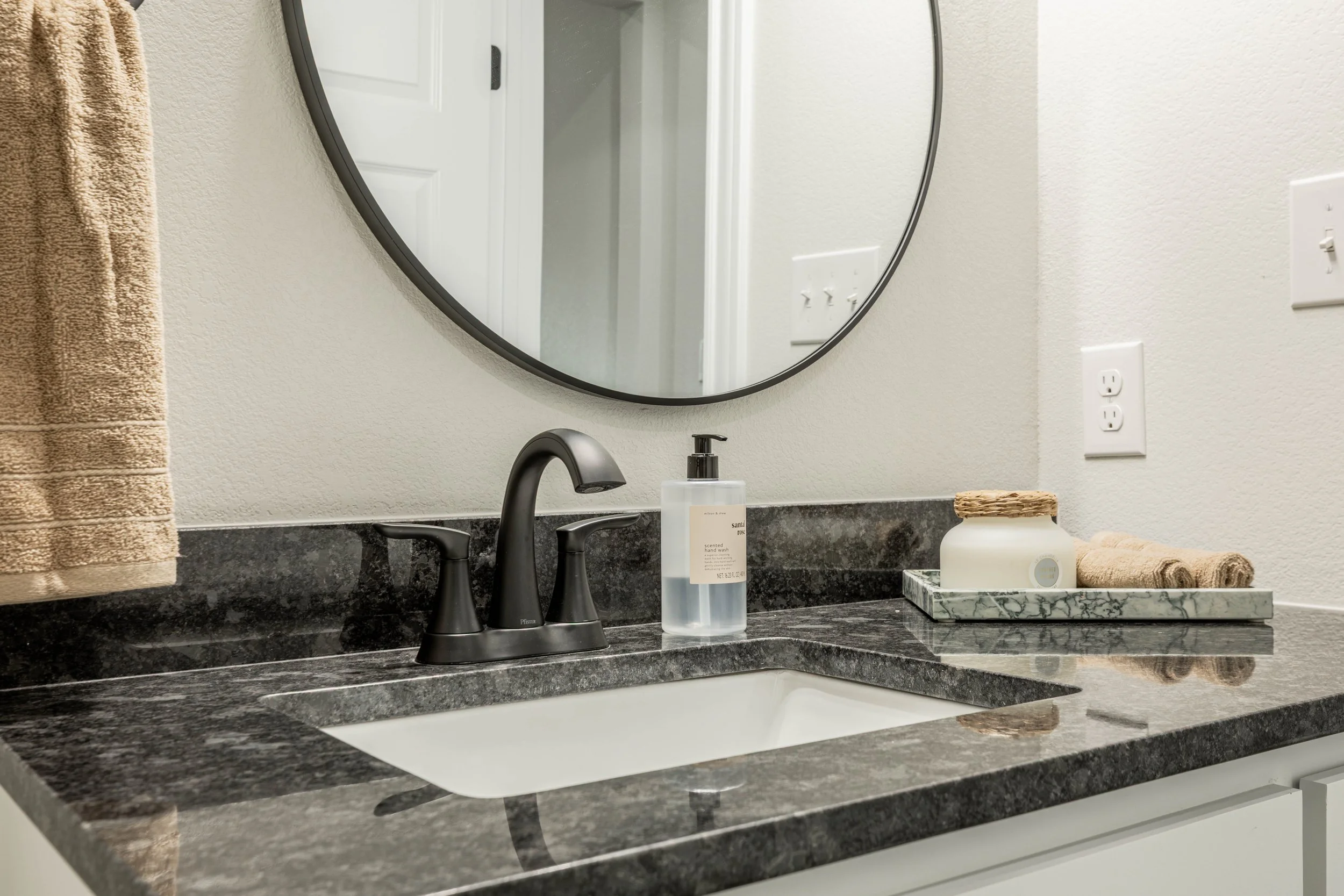 Bathroom vanity with a marble countertop, a black faucet, and a toothbrush holder. There is a mirror, a soap dispenser, and rolled towels on a tray, with a towel hanging on the left side. There are electrical outlets and a light switch on the wall.