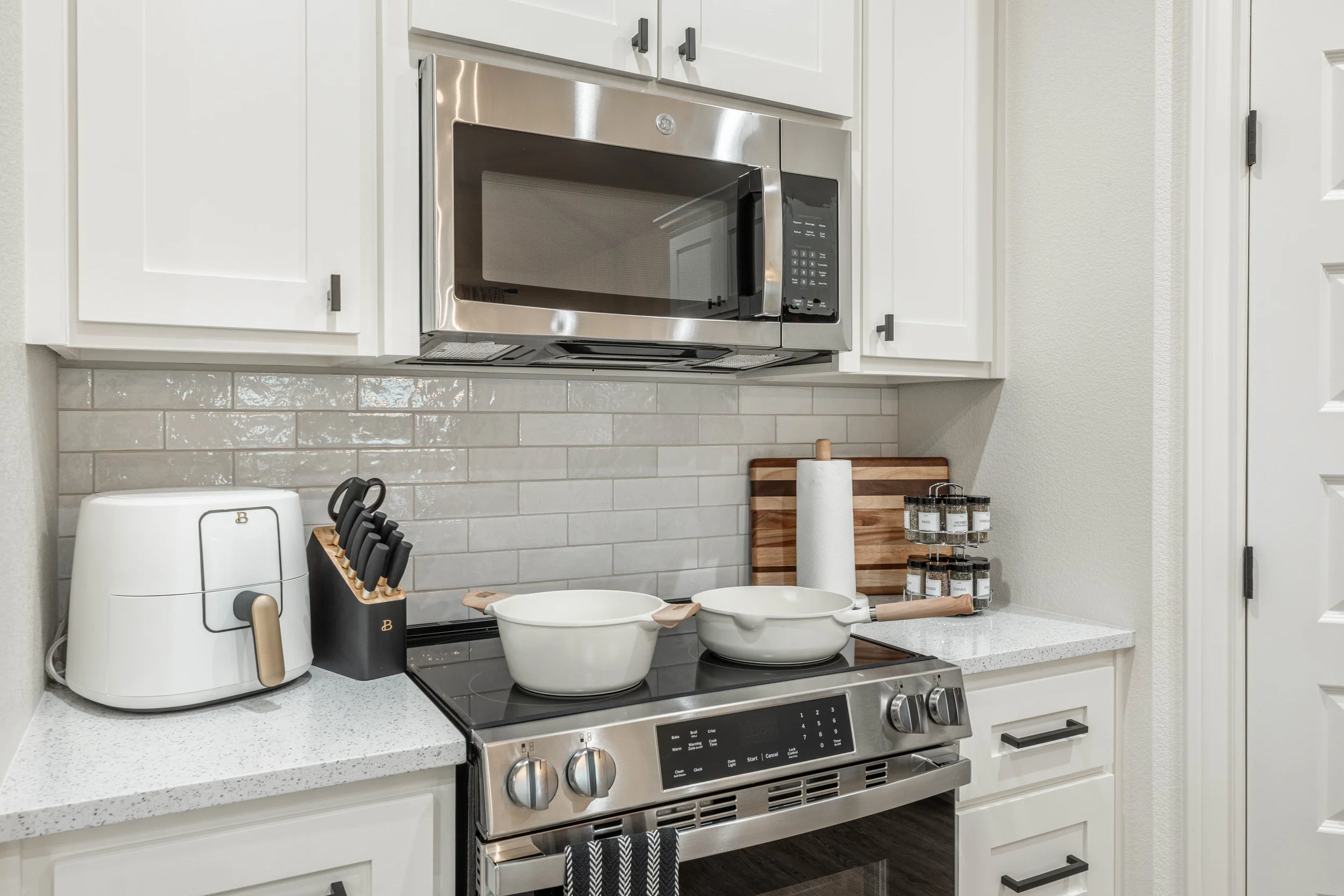 A modern kitchen with white cabinets, a stainless steel microwave above a stove, and various kitchen utensils and containers on the countertop.