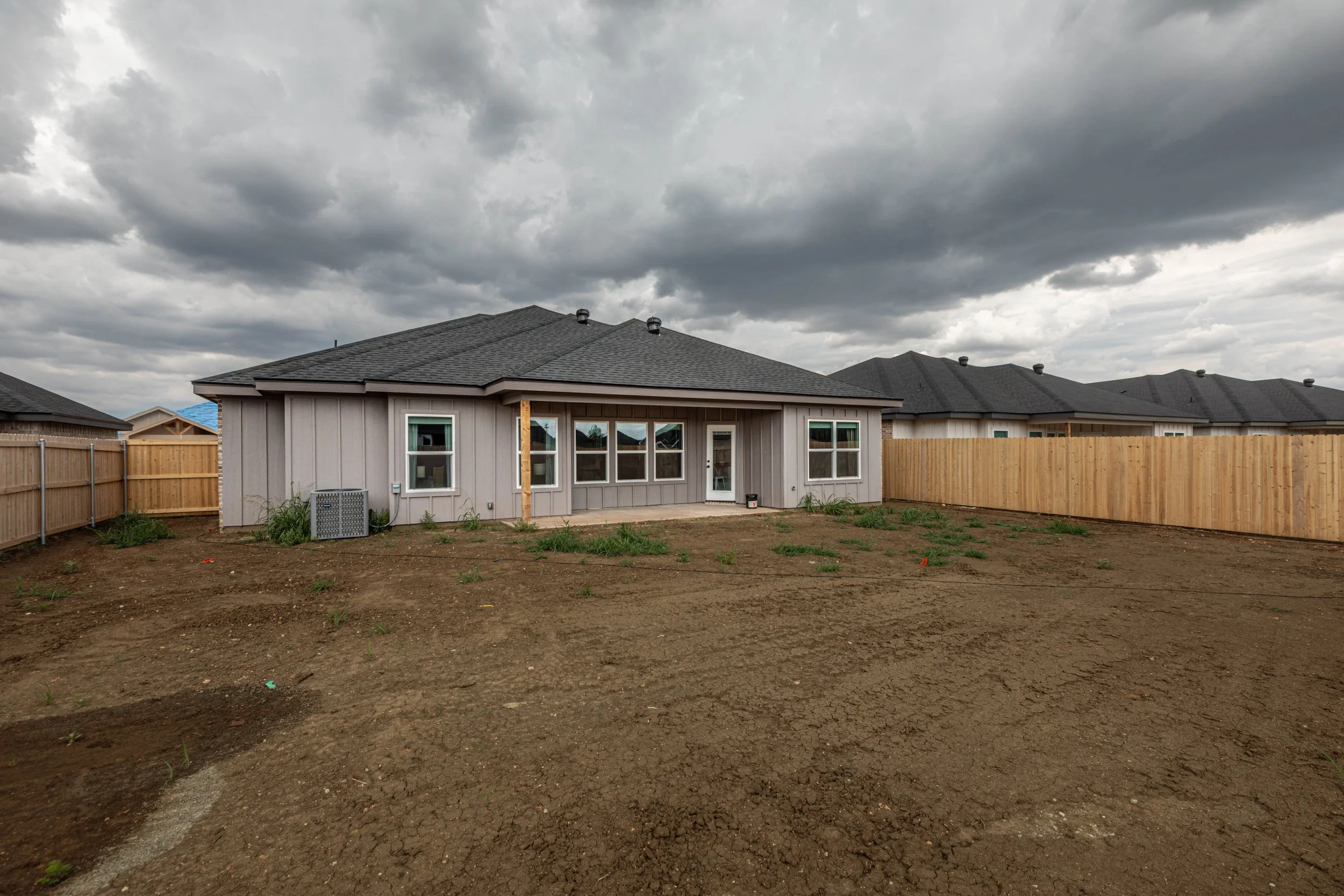 Backyard of a house with a dirt patio, wooden fencing, and cloudy sky overhead.