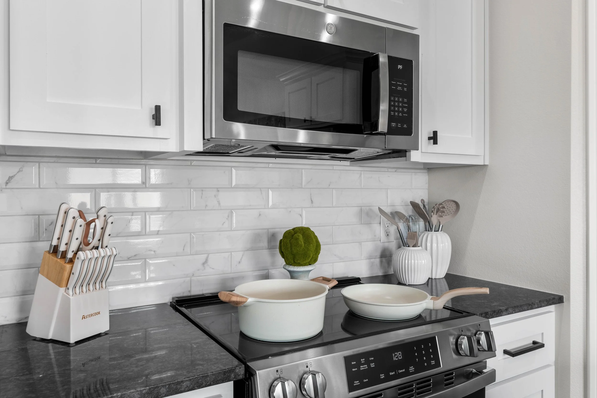 A modern kitchen with white cabinets, a marble tile backsplash, and stainless steel appliances including a microwave and a stove. There are kitchen utensils, a knife block, and white decorative vases on the counter.