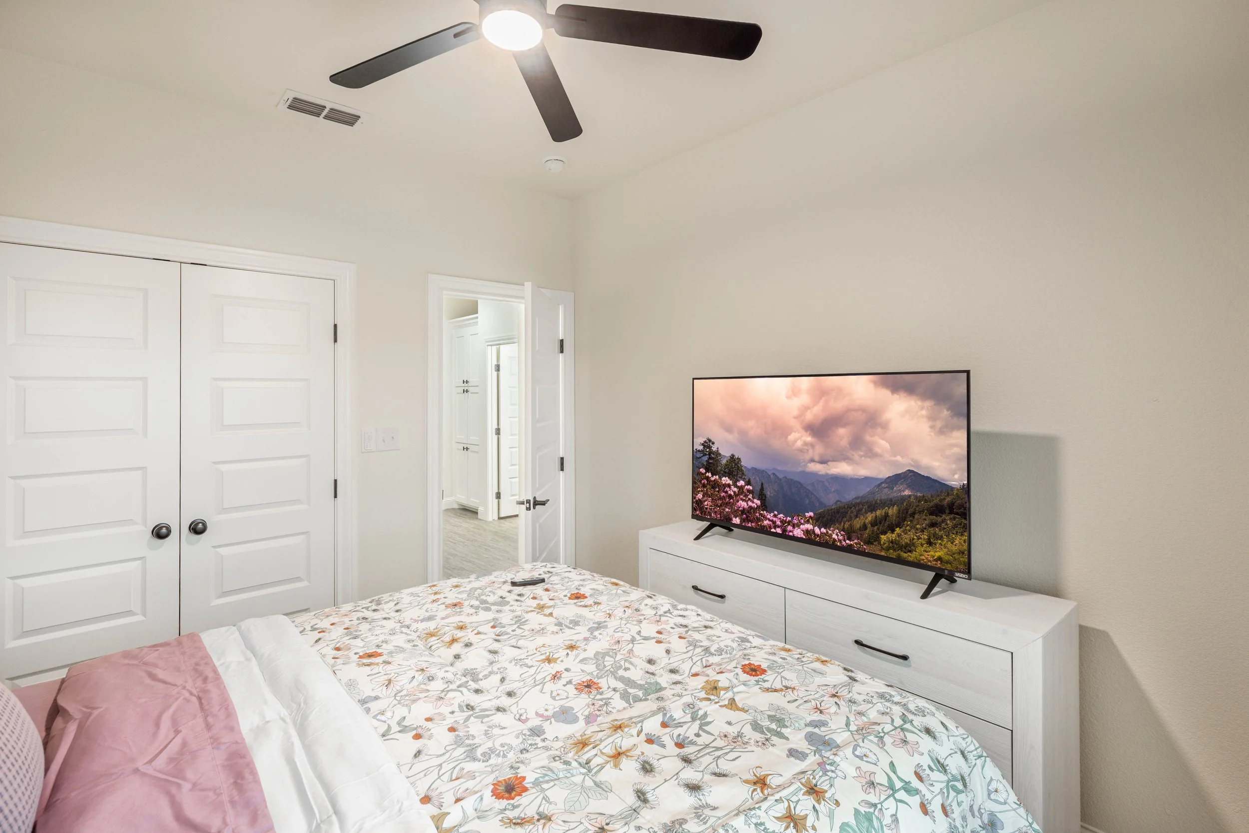 A bedroom with a ceiling fan, a flat-screen TV on a white dresser, a closed white closet door, an open door leading to another room, and a bed with floral and pink bedding.
