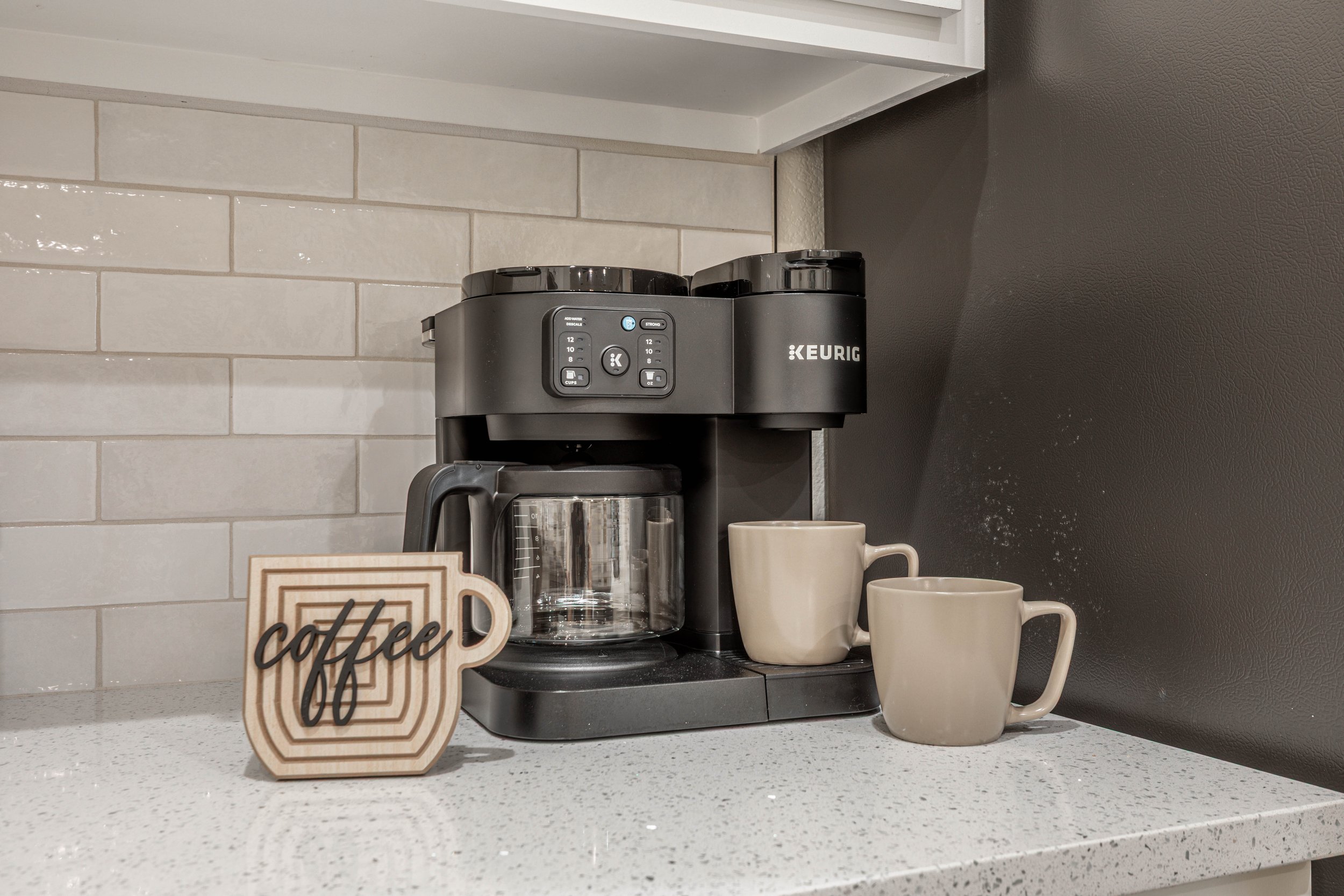 A black Keurig coffee maker on a white countertop with two white coffee mugs and a decorative coffee sign in front of it, against a wall with light gray subway tiles and a dark gray painted section.