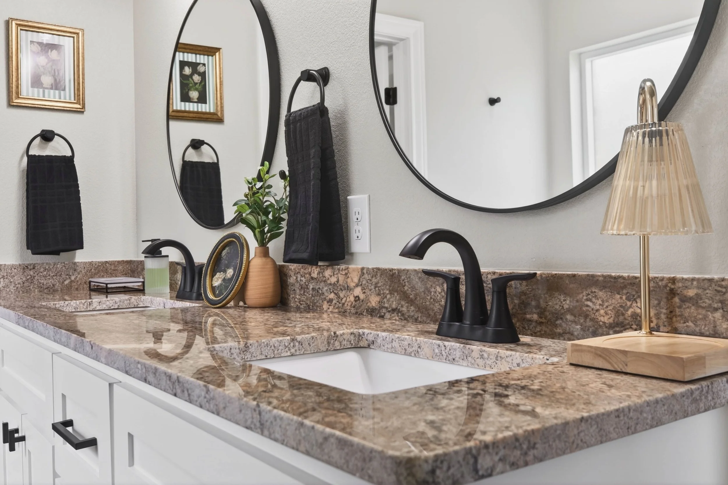 Bathroom vanity with a granite countertop, mirror, black faucet, black towel, potted plant, framed artwork, lamp, and electrical outlet.