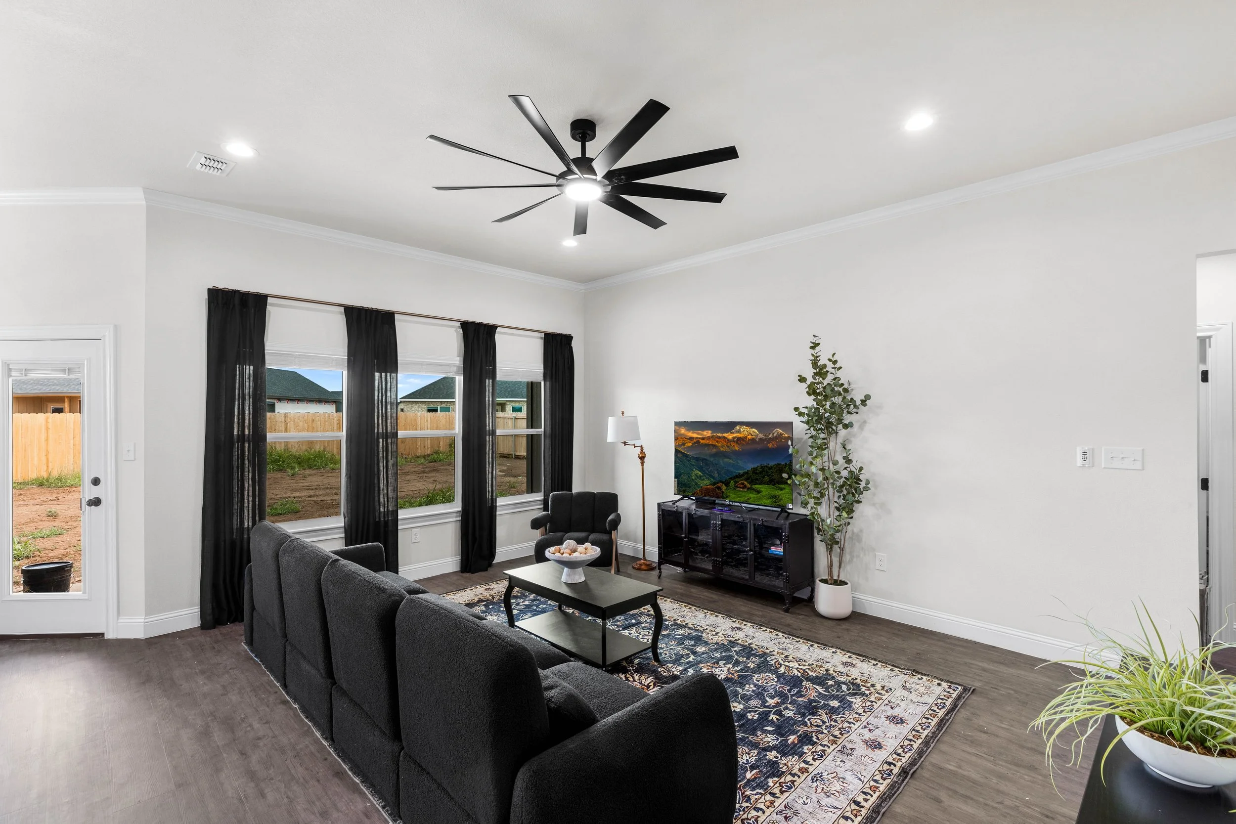 Living room with black sofa, coffee table with decorative bowl, entertainment center with TV, potted plant, and large windows with black curtains.