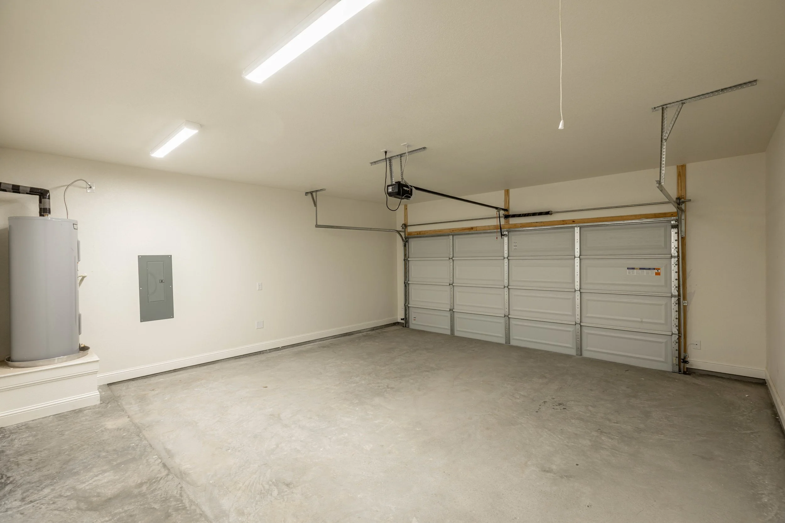 Empty garage with concrete floor, white walls, overhead lighting, a water heater on the left, garage door opener on the ceiling, and a closed garage door.