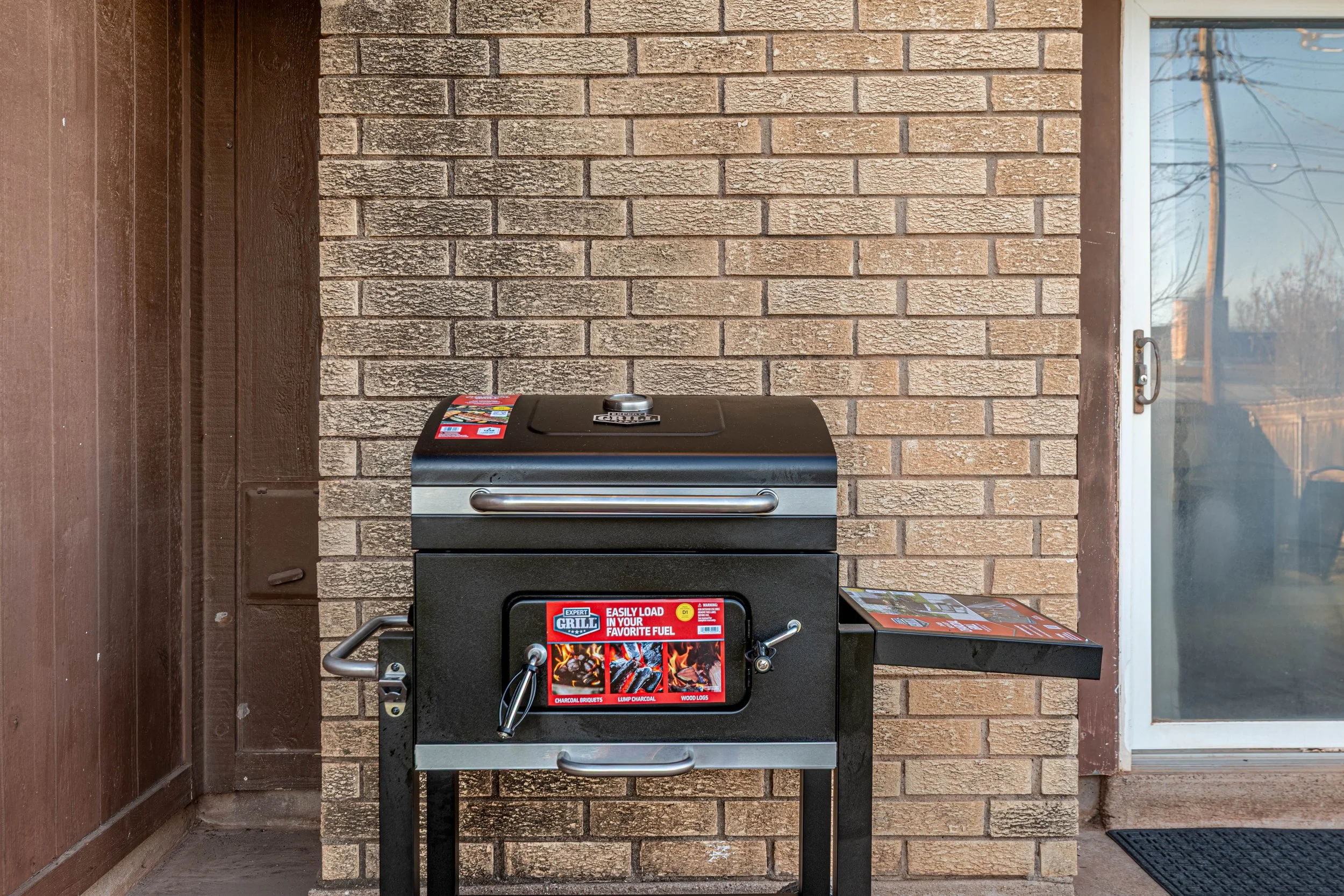 A black outdoor gas grill with a red label on the front door, positioned against a brick wall and wooden siding on the left, with a window on the right showing trees and power lines outside.
