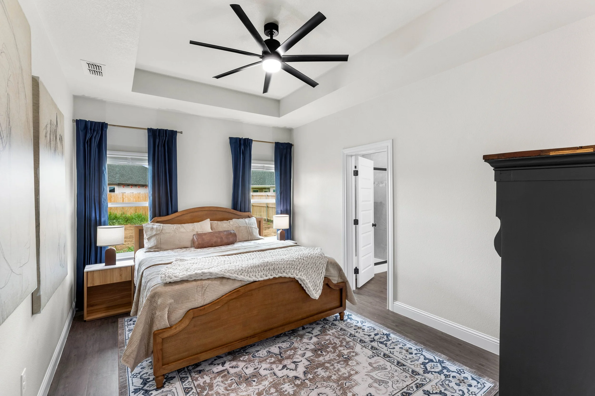 A bedroom with a wooden bed, beige bedding, navy blue curtains, two bedside lamps, a ceiling fan, and an area rug.