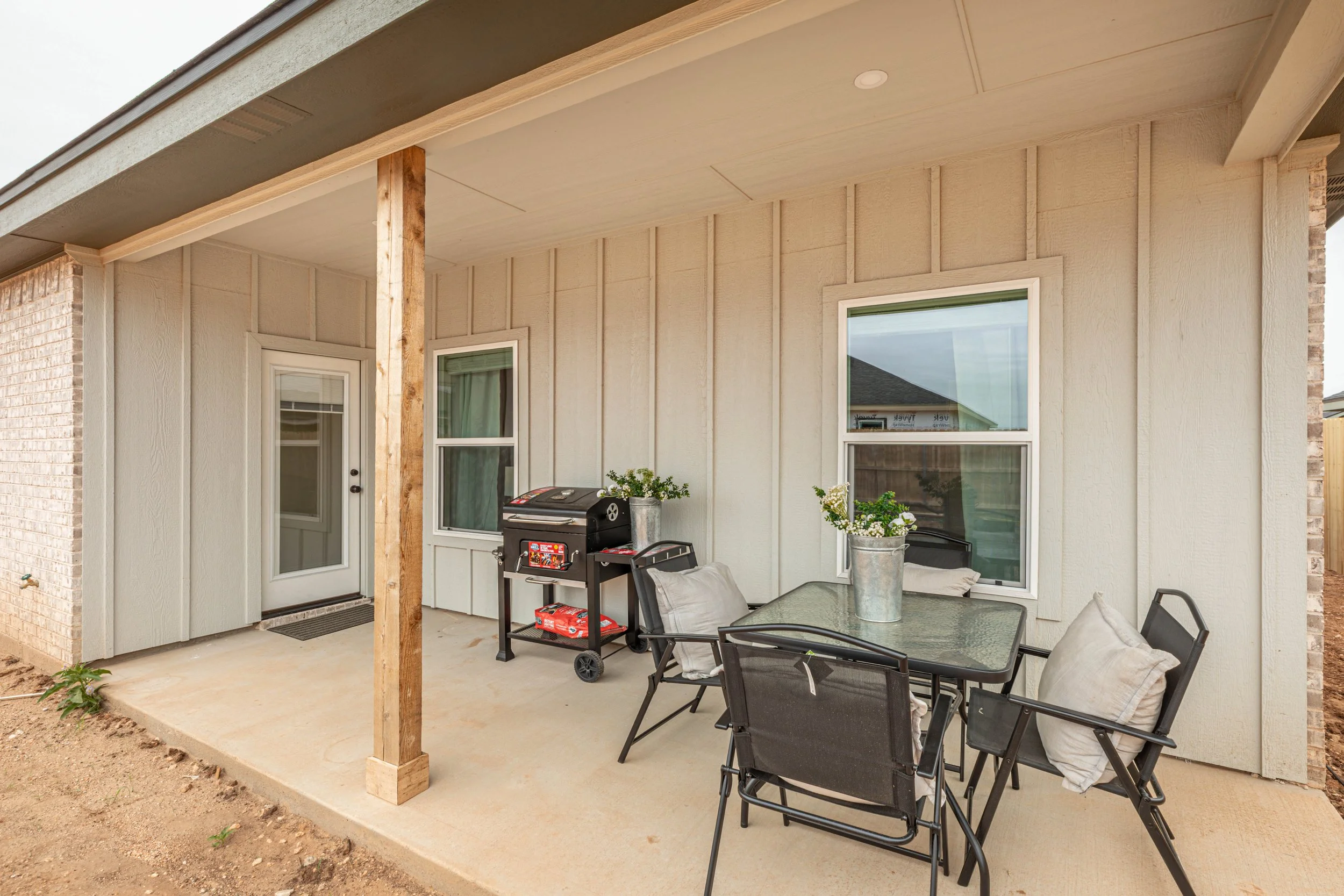 Outdoor patio area with a concrete floor, black metal furniture including a glass table and chairs with cushions, a barbecue grill, and potted plants. The patio is partially covered with a ceiling, supported by a wooden post, and has a door and two w