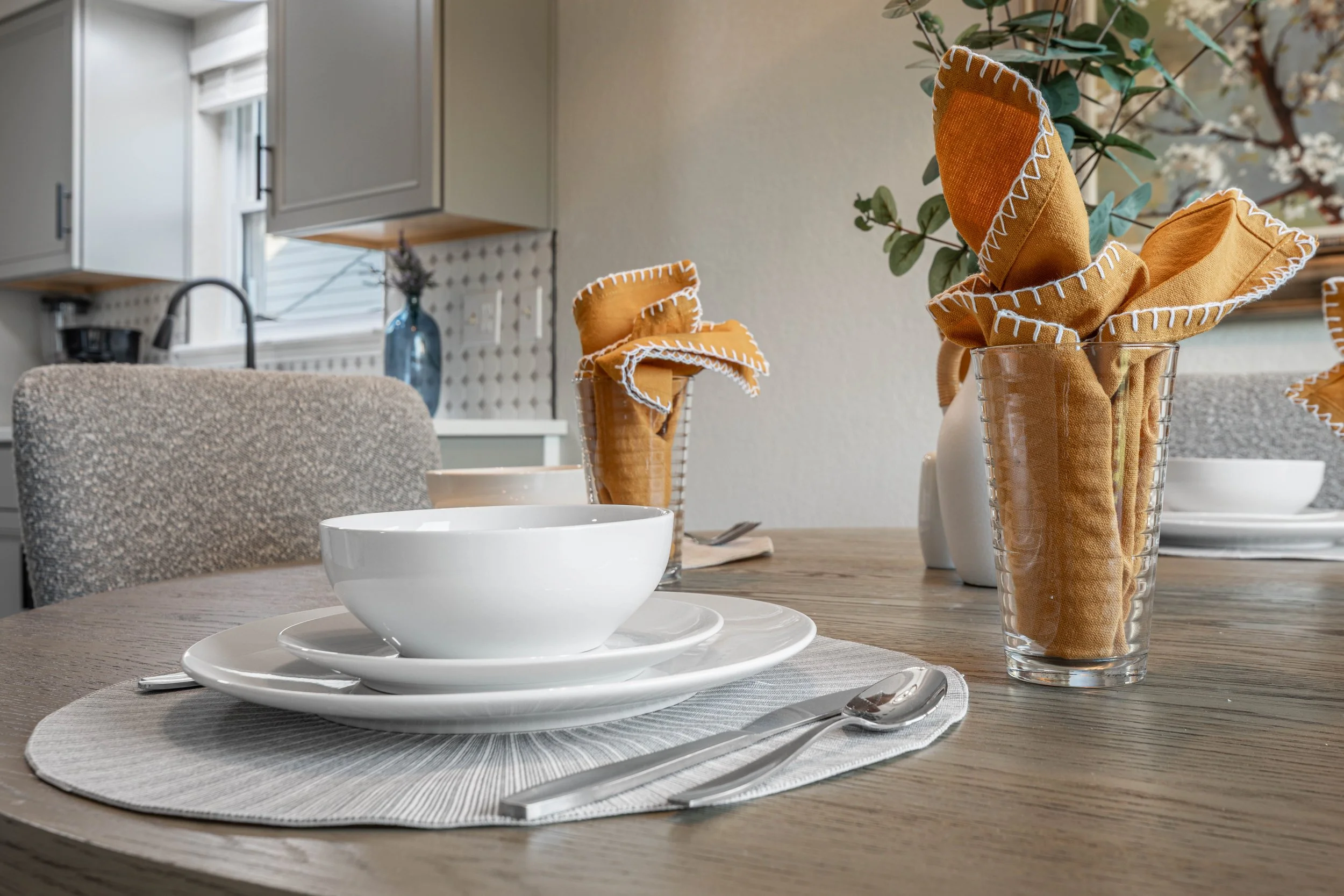 A dining table set with white bowls and plates, a spoon, and cloth napkins with orange and white stitching in a glass holder, in a bright kitchen.