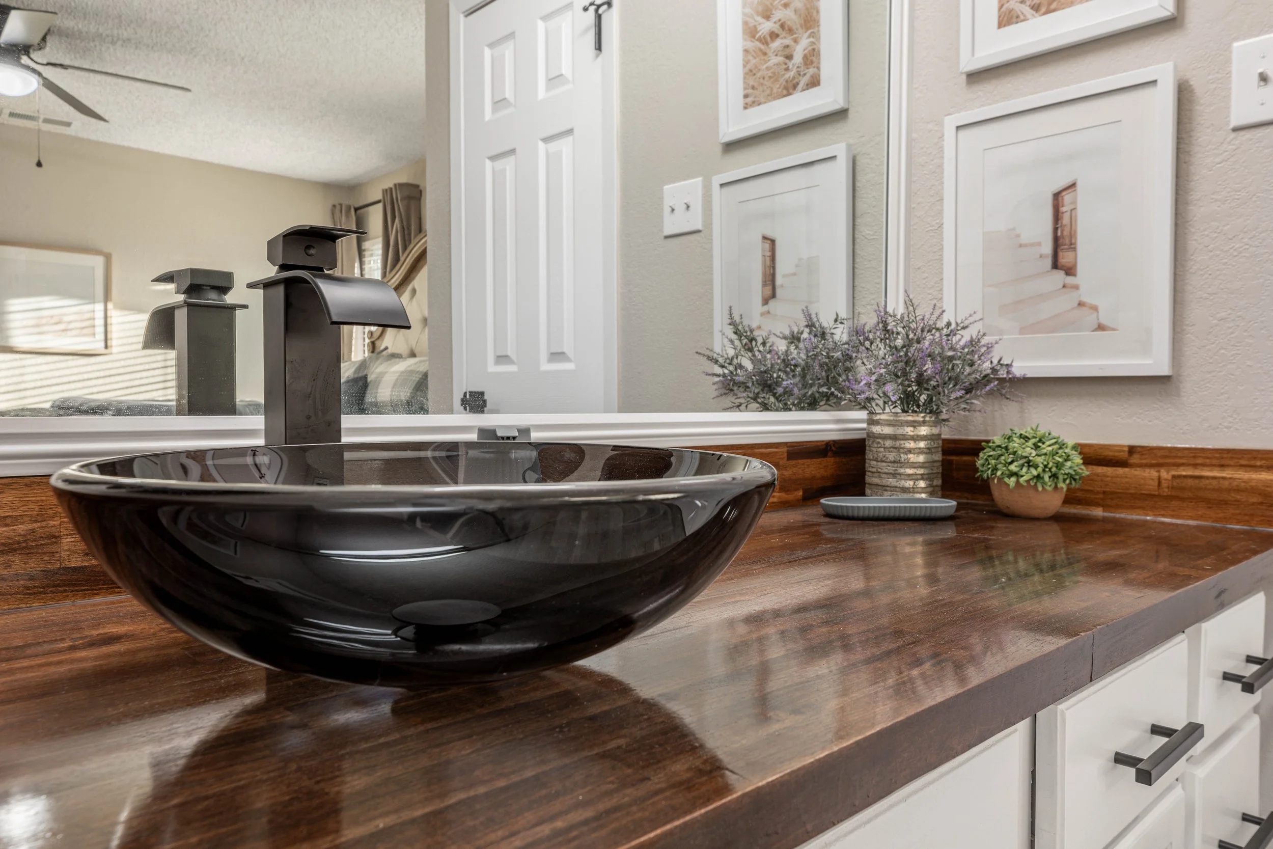Indoor kitchen with a black bowl on a wooden counter, decorative framed pictures on the wall, and a vase with purple flowers.