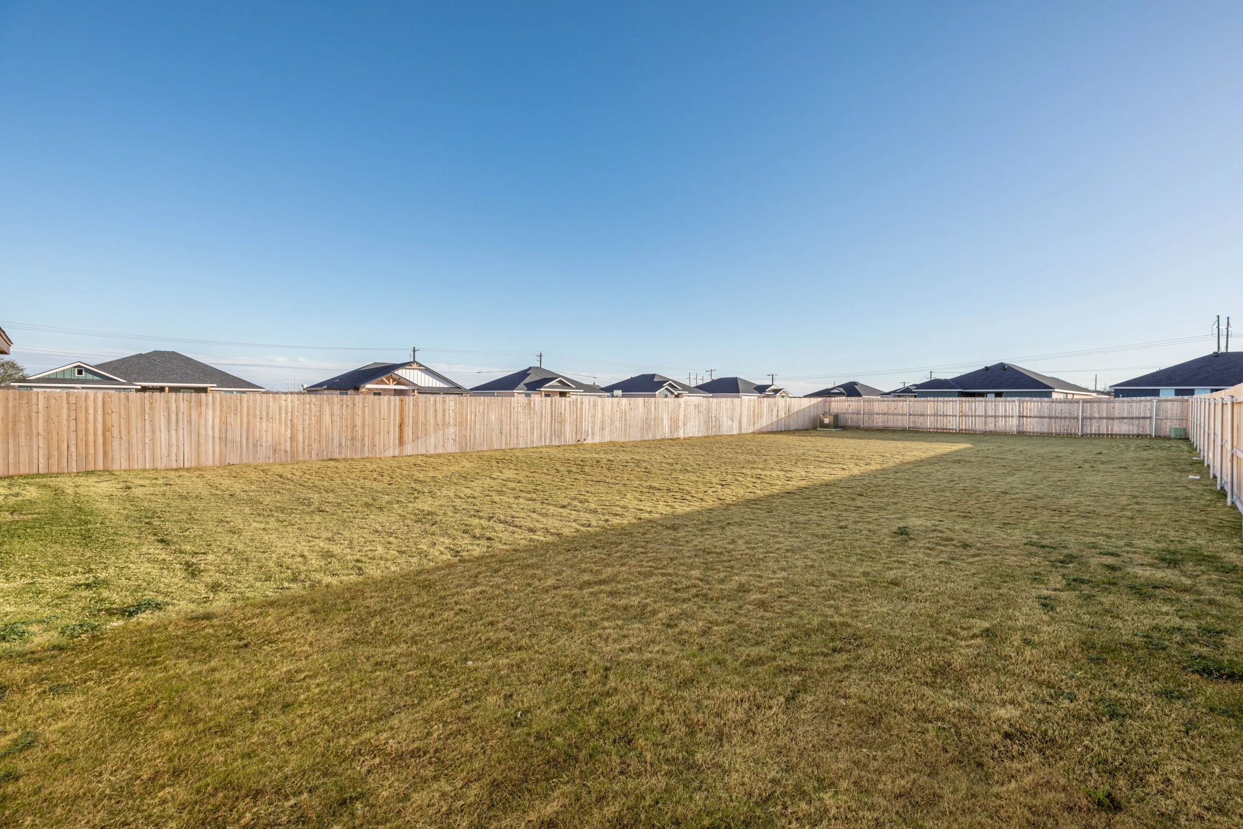 A backyard with a green lawn, wooden fence, and neighboring houses with gray rooftops under a clear blue sky.
