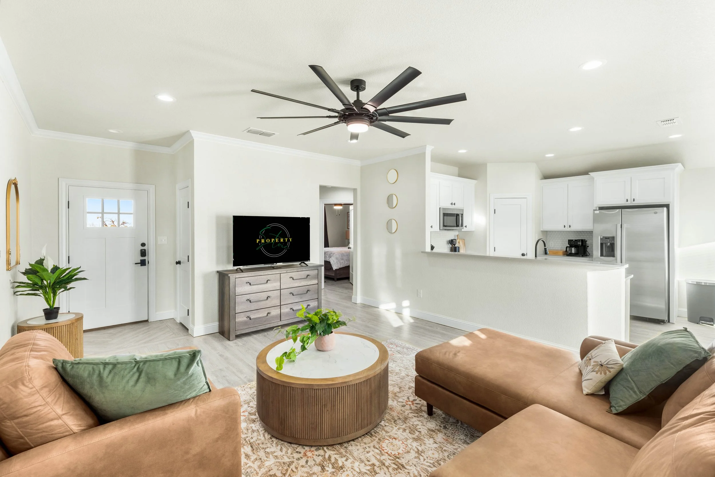 A cozy living room with brown leather sofas, green and patterned pillows, round wooden coffee table with plants, white walls, cream-colored carpet, a TV on a wooden stand, and a kitchen area with white cabinets, microwave, refrigerator, and black cof