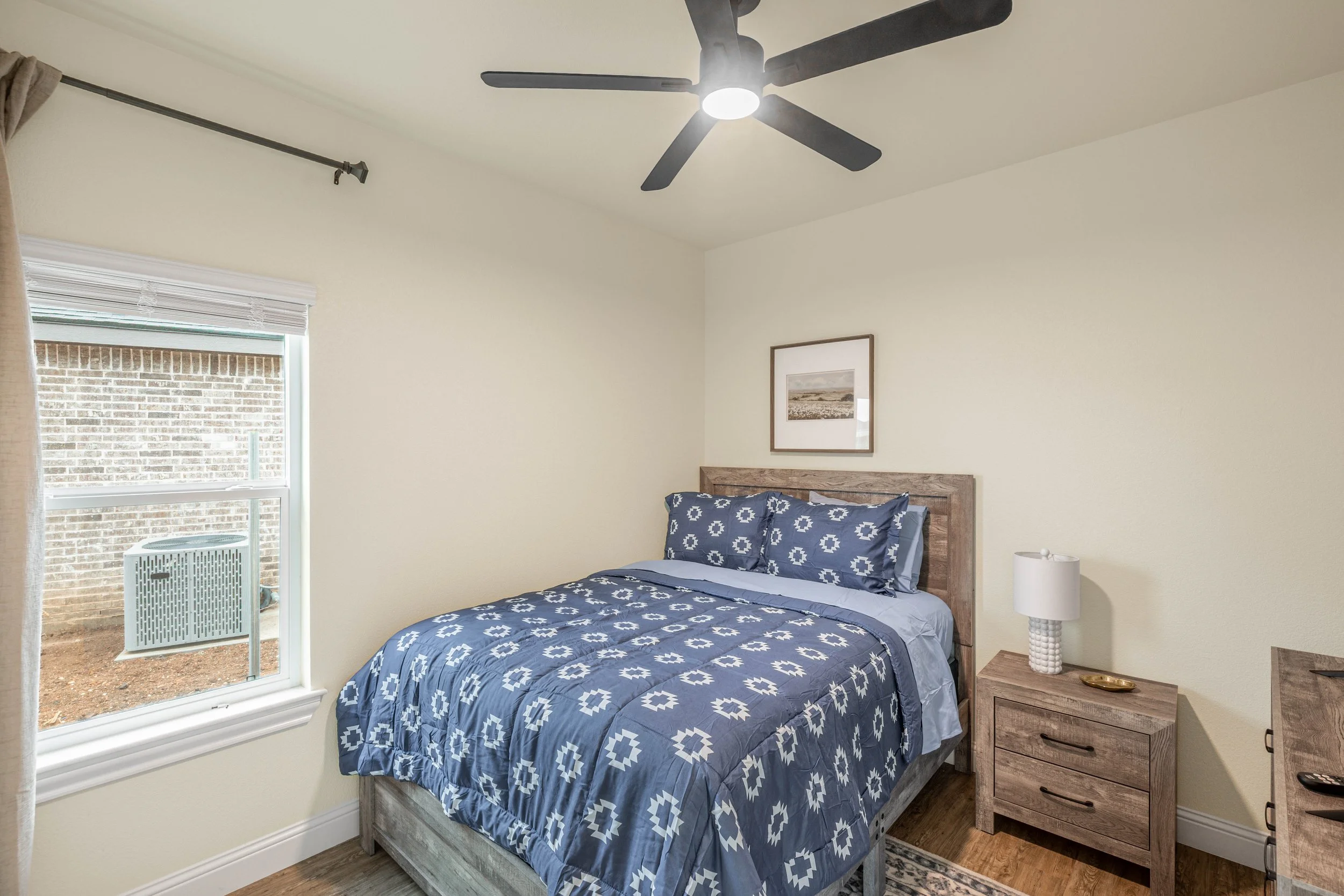 A bedroom with a bed covered in blue and white patterned bedding, a wooden bedside table with a white lamp, a framed landscape picture on the wall, a window with partially open blinds, a ceiling fan with a light, and a view outside showing a brick wa