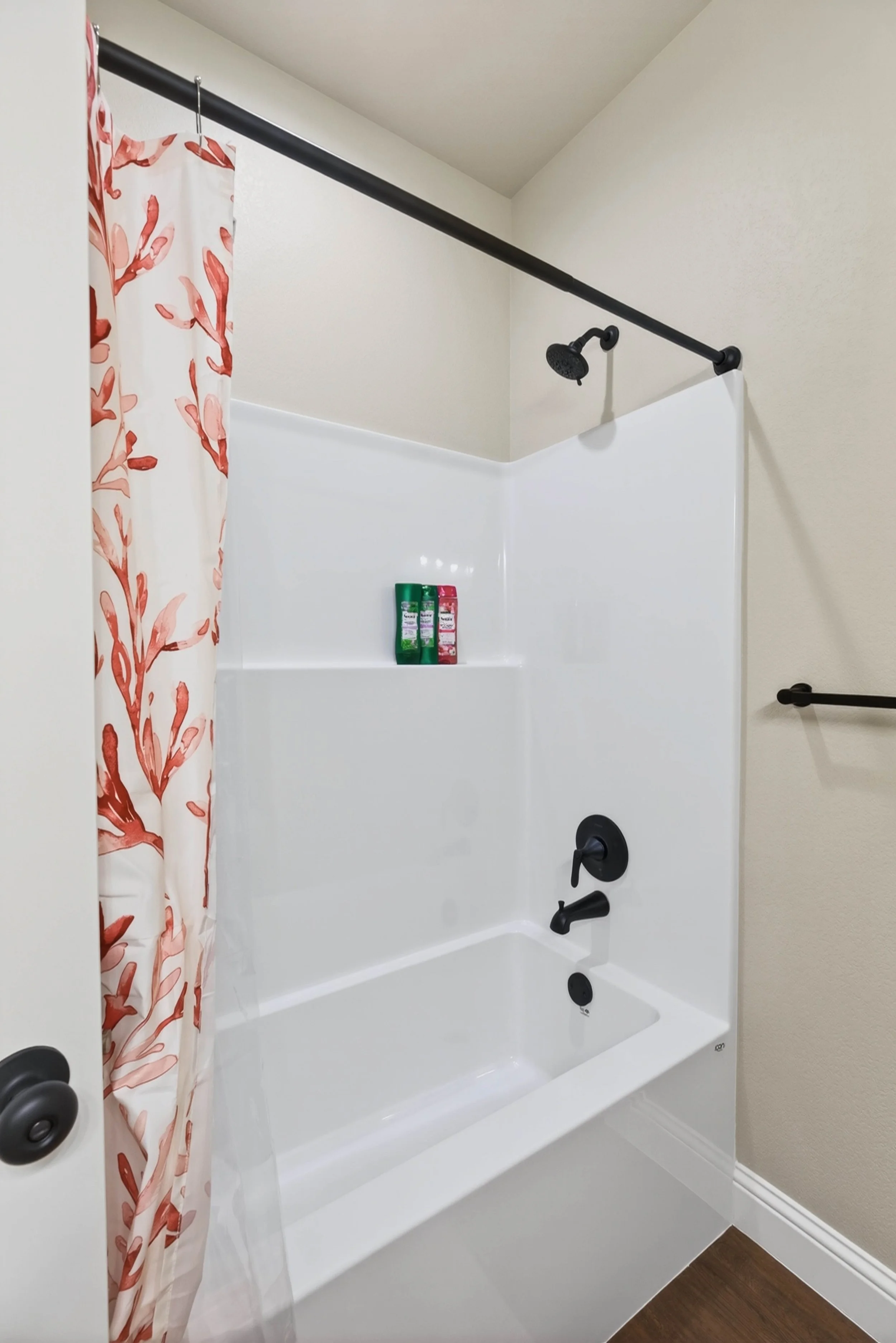 A bathroom shower with a white bathtub, a black showerhead, and black controls. There is a pink and white shower curtain with a floral pattern on the left side. Two bottles of shampoo or body wash are placed on a ledge inside the shower.