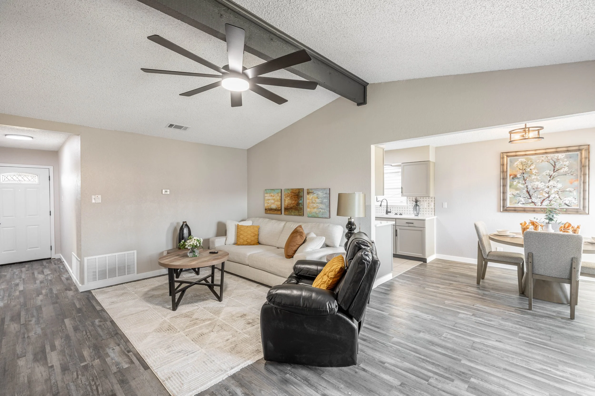 Living room with a ceiling fan, a white sofa with yellow cushions, a coffee table with plants, a black leather armchair, and a dining area with a table and chairs near the kitchen.