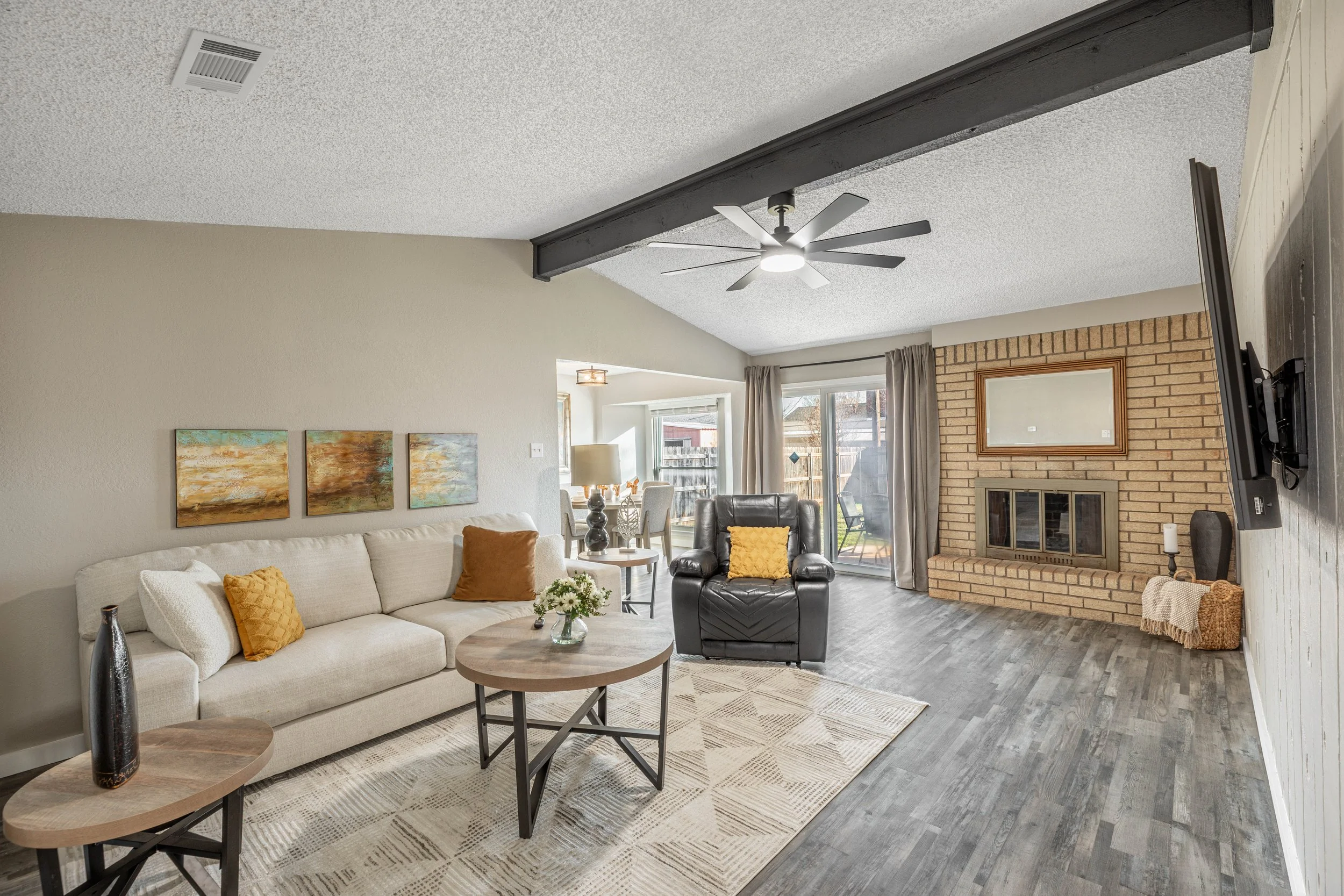 Living room with a beige sofa, yellow throw pillows, a coffee table with a vase of flowers, a black leather chair with a yellow pillow, a brick fireplace, wall-mounted television, and large windows with curtains.