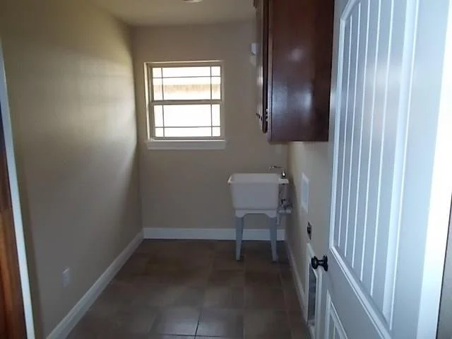 A small laundry room with beige walls, a window, a white utility sink, dark wood cabinets, and a white door.